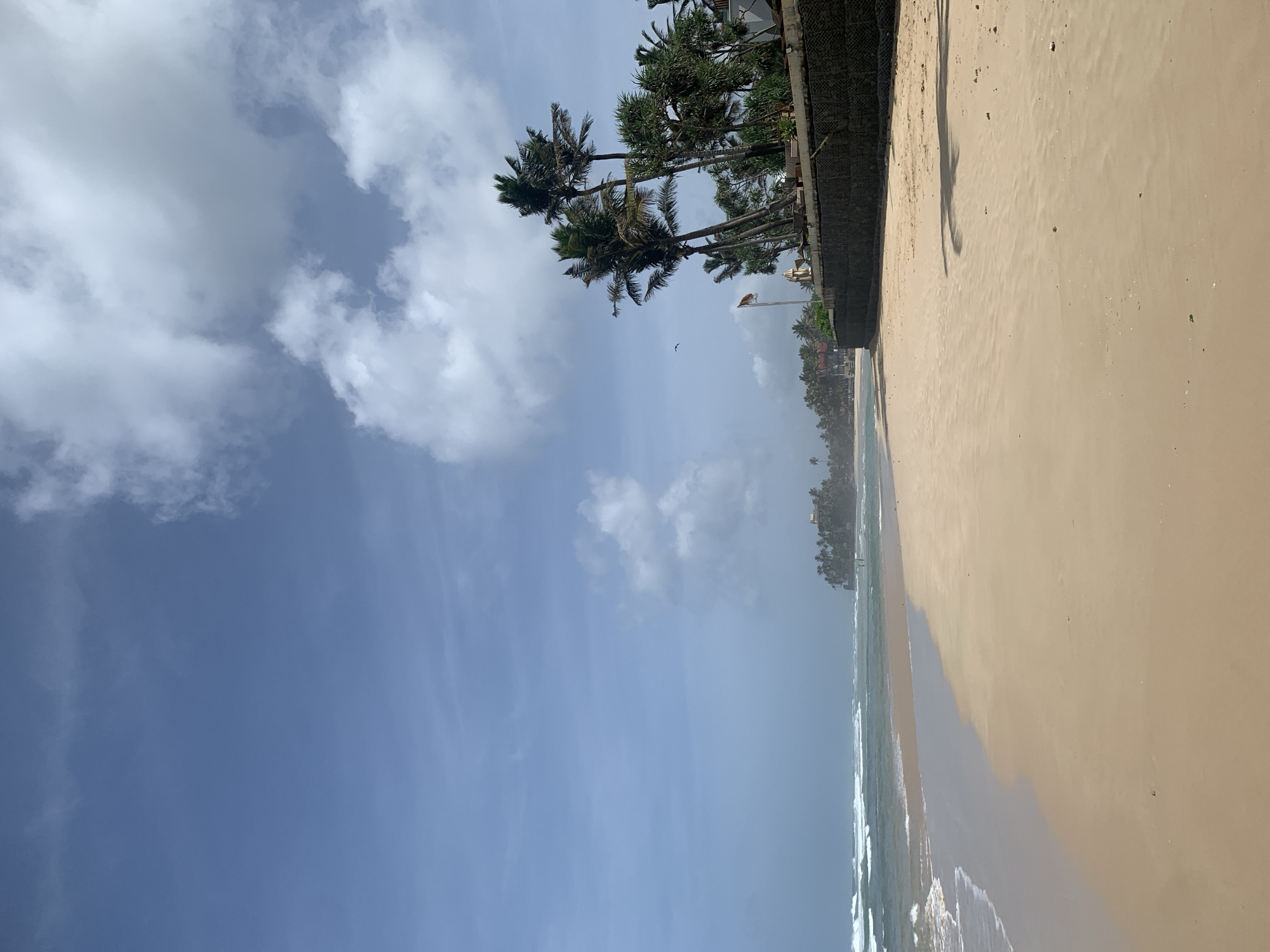 The image captures a serene beach scene in Kathaluwa, Sri Lanka, under a bright blue sky dotted with fluffy white clouds. The foreground is dominated by a wide expanse of golden sand, gently sloping towards the ocean. The waves are rolling in, creating a foamy white edge along the shoreline, and the water appears a tranquil turquoise.
To the right of the frame, a stone wall, possibly a seawall or part of a resort, runs along the edge of the beach. Several tall palm trees with lush green fronds stand behind this wall, casting long shadows on the sand. A flag, which appears to be the Sri Lankan flag, is visible flying from a flagpole near the palm trees. In the distance, more palm trees and structures suggest a developed coastal area. A few scattered individuals can be seen walking along the beach in the far distance, adding a sense of scale and human presence to the otherwise natural landscape. The overall atmosphere is peaceful and idyllic.