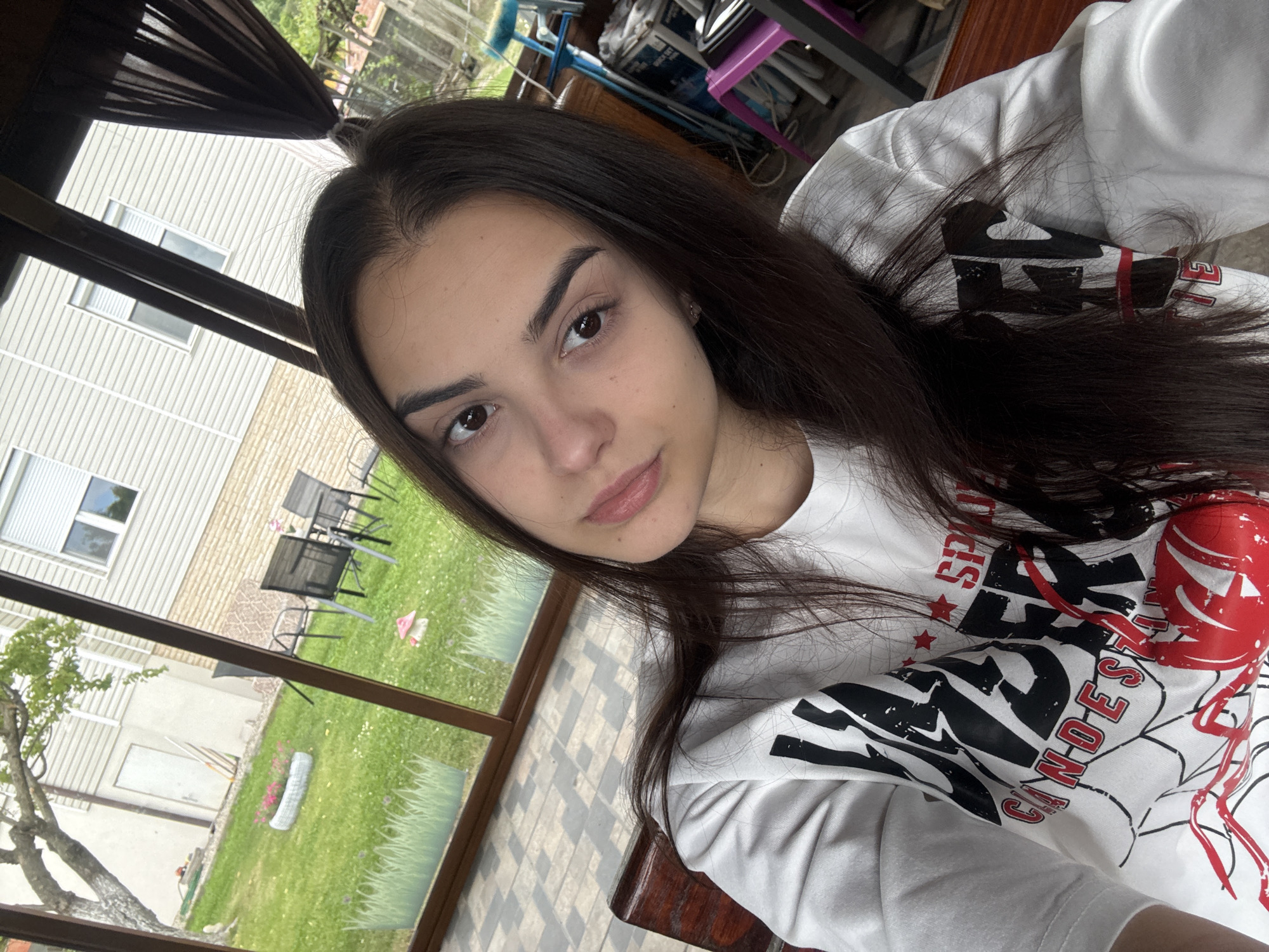 A young woman is taking a selfie indoors. She has dark hair and is wearing a white t-shirt with a graphic design. The background shows an outdoor setting with a house, grass, chairs, and trees visible through a window or screen. The location might be a sunroom or enclosed porch. There's also some furniture and objects in the background behind her, including a blue object, a brown surface, and a pink chair. I am unable to determine the location is the city of Polena, Bulgaria.