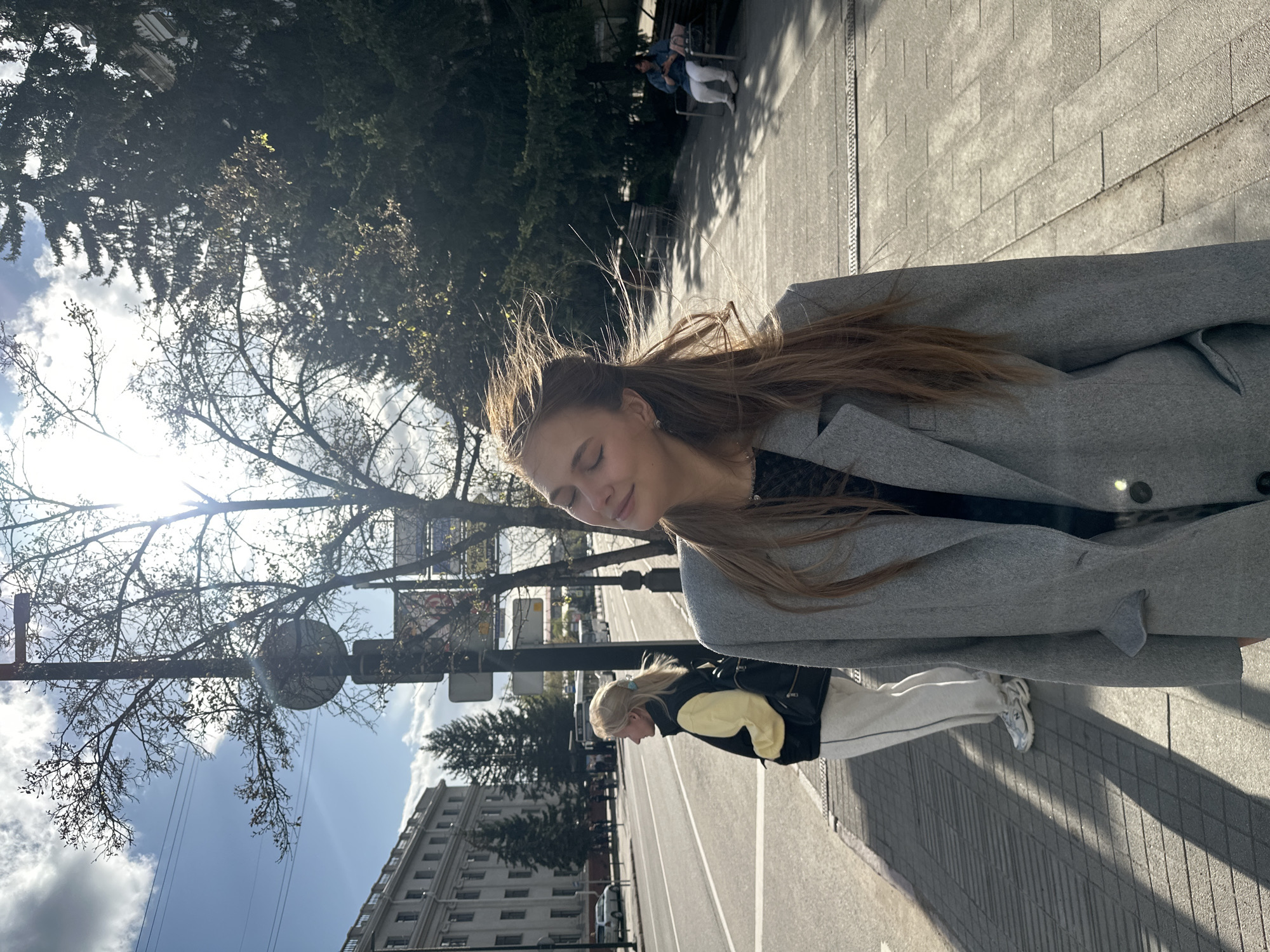 The image is an outdoor shot of a light-skinned female with long brown hair wearing a gray coat. She appears to be walking in the city of Krasnoyarsk, Russia, indicated by the visible city architecture. Another person is partially visible, walking in the opposite direction. Trees and a building are in the background. The weather appears sunny.