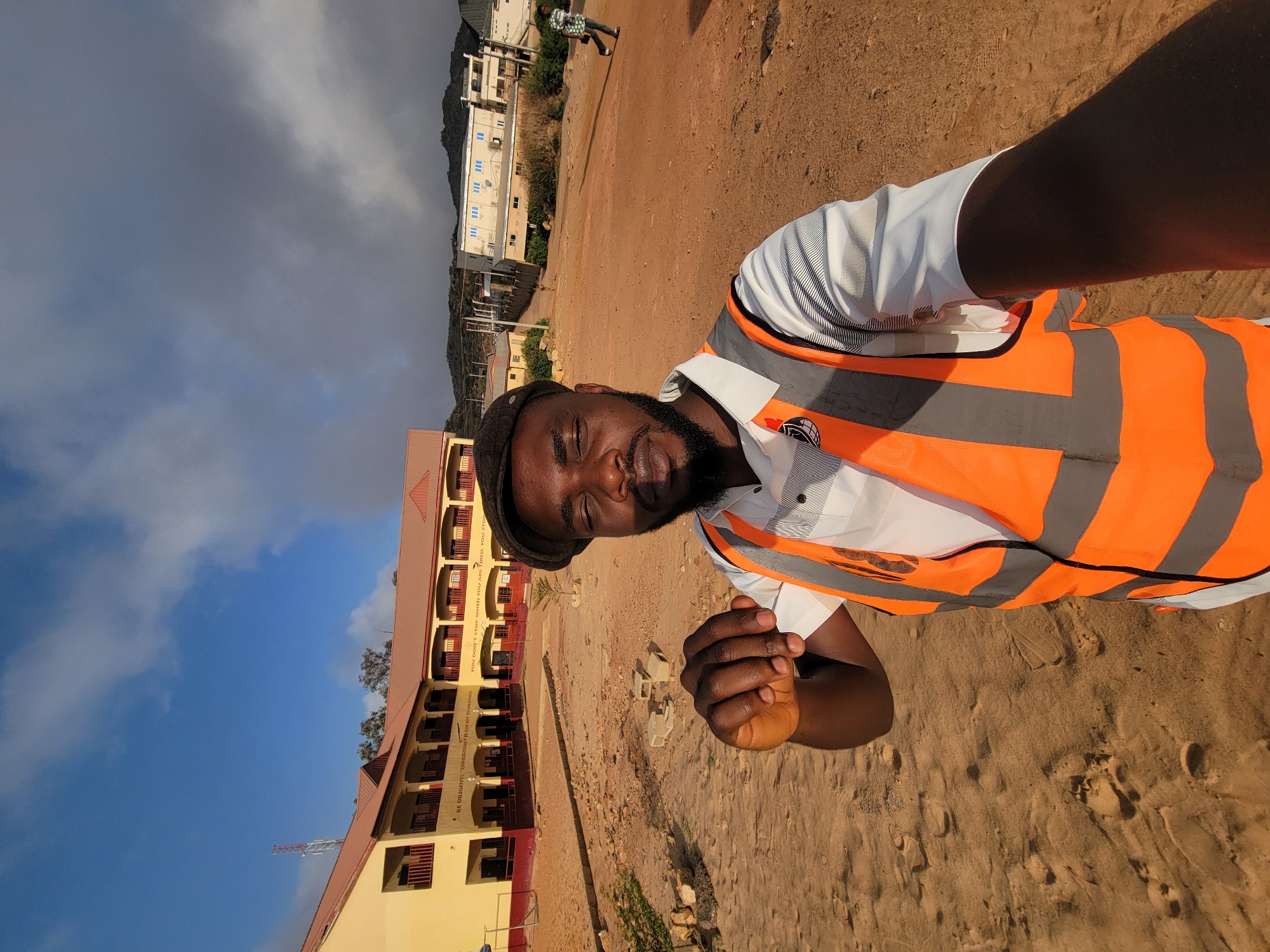 A medium shot shows a man wearing a white shirt and an orange and gray reflective safety vest, standing on a sandy patch of land, presumed to be near Gura Top, Nigeria, based on the prompt. The man, who appears to be Black, is facing the camera with closed eyes. He is wearing a black cap. In the background, there is a multi-story yellow building with red roof and visible inscription on the side of the building. Other structures are visible in the background along with a person walking. The sky is partly cloudy with a mix of blue and gray.