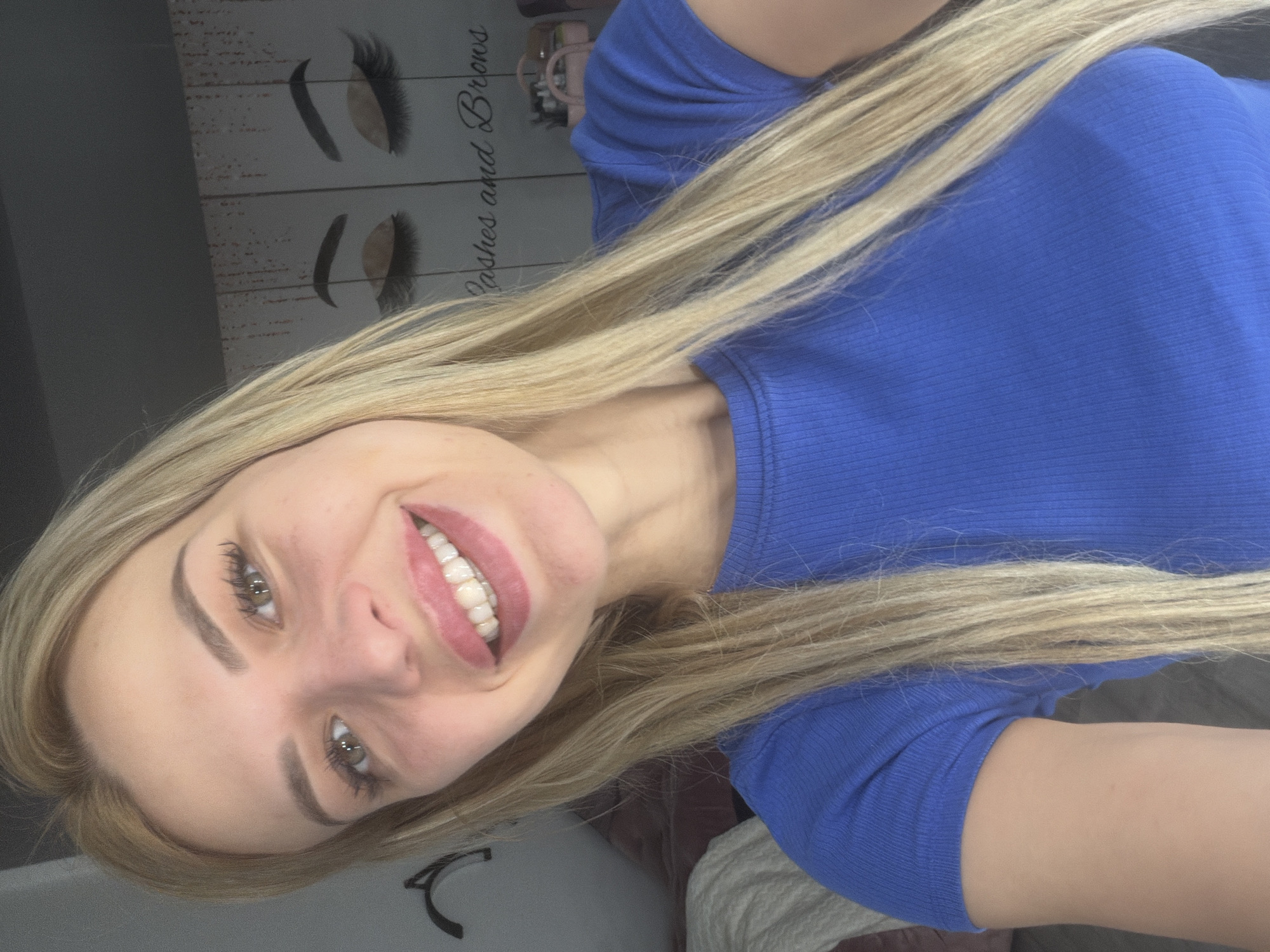 A blonde woman smiles at the camera in a close-up shot. She is wearing a blue, ribbed t-shirt. Her hair is long and blonde, falling around her shoulders. In the background, a wall decoration features drawings of eyelashes and eyebrows, with the text "Lashes and Brows." The scene appears to be indoors, possibly in a beauty salon or a home environment. There is no indication of the location being in Kyiv, Ukraine.