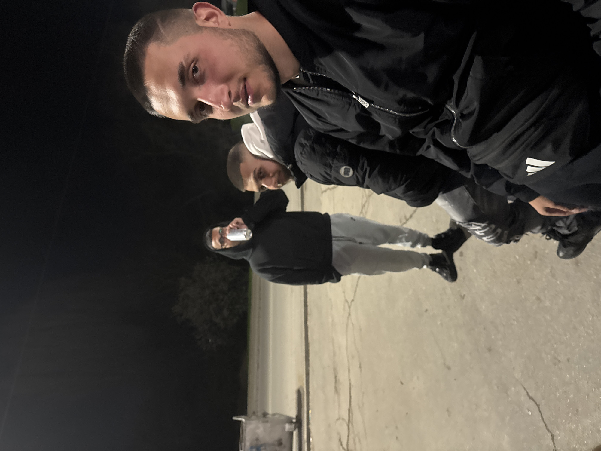 Three young men are standing outside at night. The ground appears to be concrete. The man in the front is wearing a black jacket and has short, dark hair. The second man is wearing a white hoodie underneath a black jacket. The third man is standing further back, wearing a black hoodie, grey sweatpants, and glasses. He is holding a can to his mouth. There is a trash receptacle on wheels visible in the lower-left corner. The location appears to be Topoli, Bulgaria.