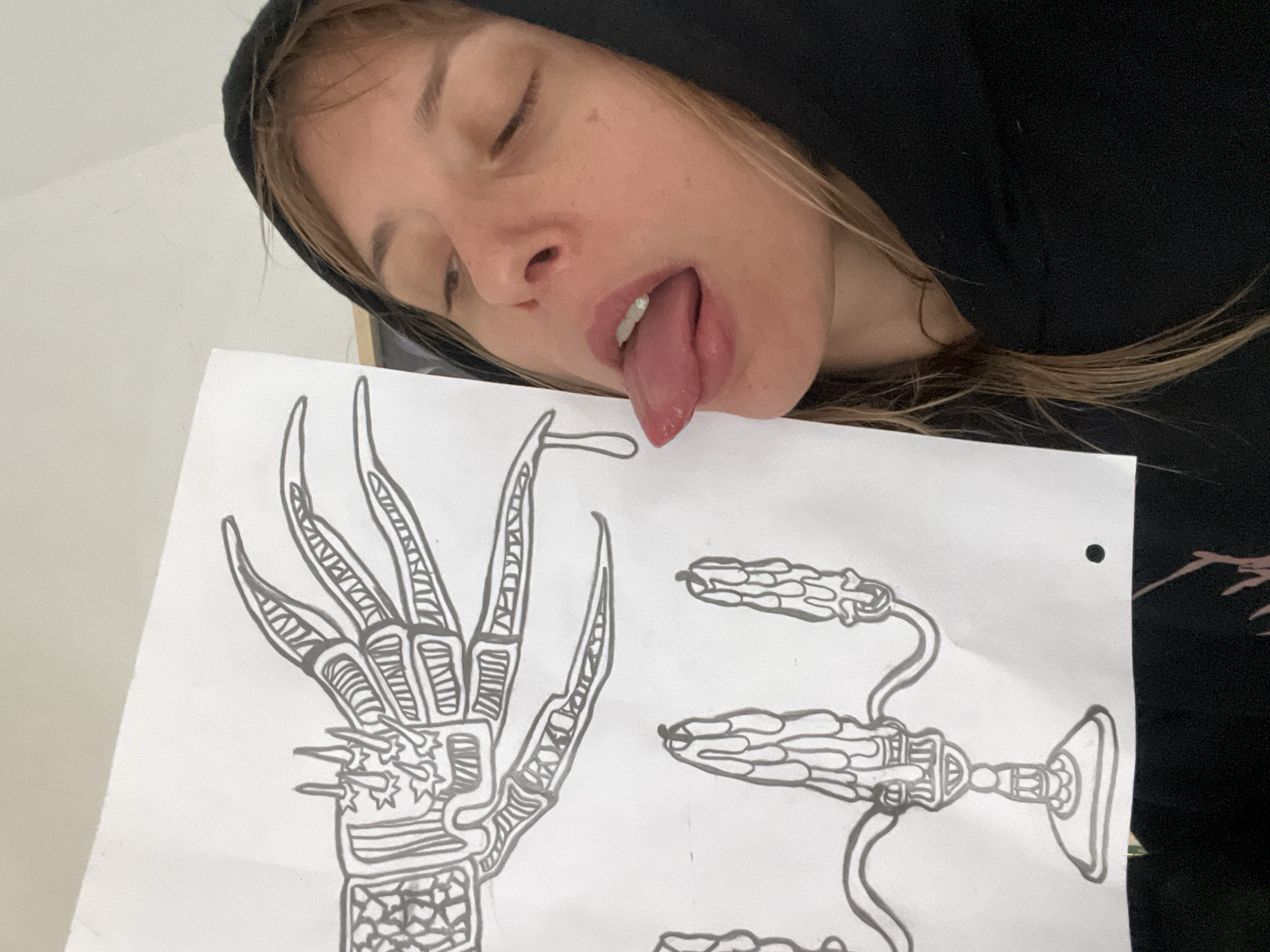 A fair-skinned person with blond hair, wearing a black hooded sweatshirt, is holding a drawing and sticking their tongue out. The drawing appears to be a black and white illustration of stylized claws and a goblet-like object connected to curved lines. The background is a plain, light-colored wall. There is no evidence to suggest the location is Buenos Aires, Argentina.