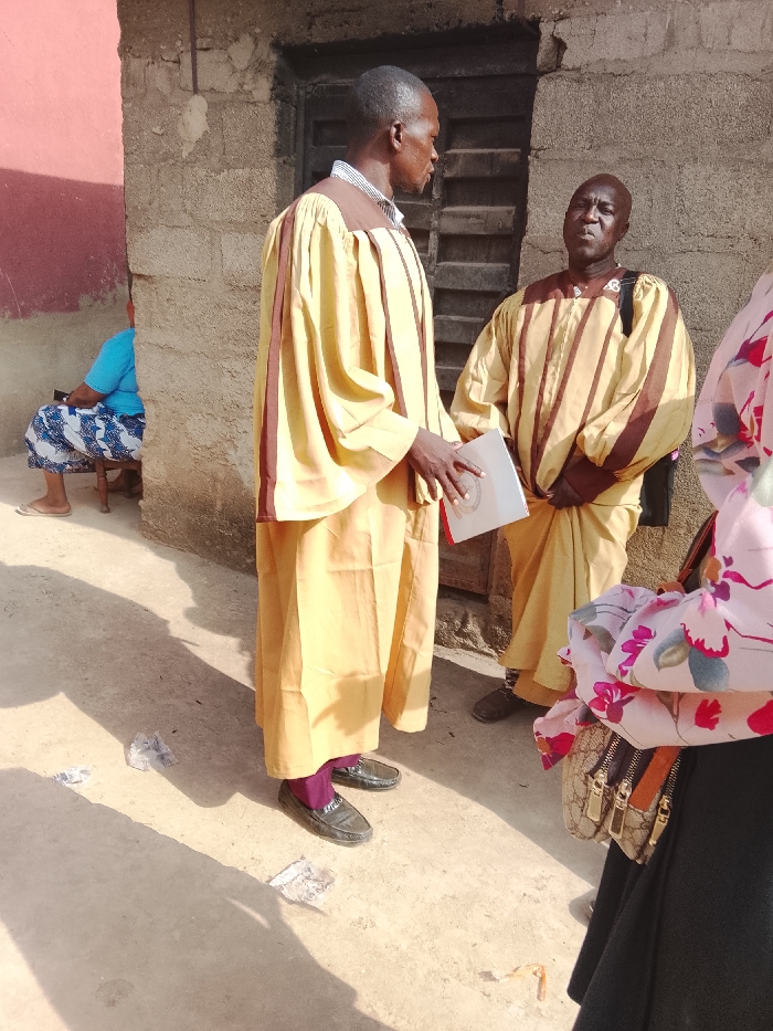 The scene depicts an outdoor gathering in Ube, Nigeria, under bright daylight conditions. Two adult males stand centrally, appearing to converse. The taller male on the left, facing right in profile, wears a long-sleeved yellow gown with brown vertical stripes and brown shoulder accents over a collared shirt. He holds a white booklet or document in his left hand. He wears black loafers and purple socks. The second male, to his right, faces forward, wearing a similar long-sleeved yellow gown with brown horizontal and vertical stripes, and a dark bag strap visible over his left shoulder. He wears dark shoes.
In the background, a rough, light grey concrete block wall is visible behind the two central males, with a dark wooden door or shuttered window recessed into it. To the left, a section of wall is painted a faded reddish-brown. A third individual, partially visible on the far left, is seated on a wooden stool, wearing a blue short-sleeved top and a blue and white patterned skirt, with light-colored sandals. A fourth individual, partially visible on the far right, wears a black skirt and a light-colored top with a pink and green floral pattern, carrying a light brown handbag with a patterned fabric and gold-colored clasps.
The ground is a light brown, possibly packed earth or concrete, showing shadows consistent with direct sunlight. Several pieces of crumpled white debris are scattered on the ground near the feet of the central figures.