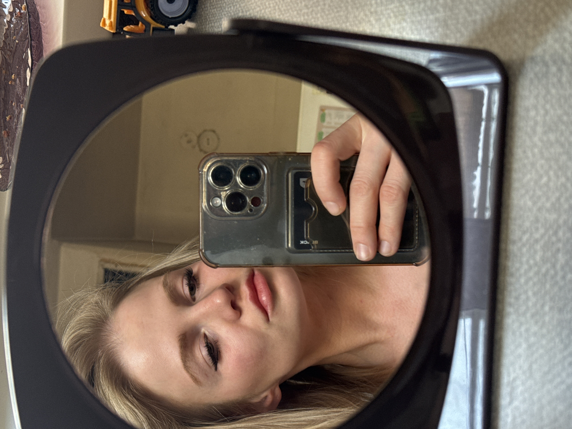 A woman is taking a selfie in a mirror. The mirror is black and oval-shaped. The woman is holding a phone with a clear case and several lenses. The phone has a card holder on the back. The woman has blonde hair and is wearing makeup. The background is a wall with peeling paint and possibly a toy vehicle on top. The location is not discernable from the image and it cannot be determined that it is in Saint Petersburg, Russia.