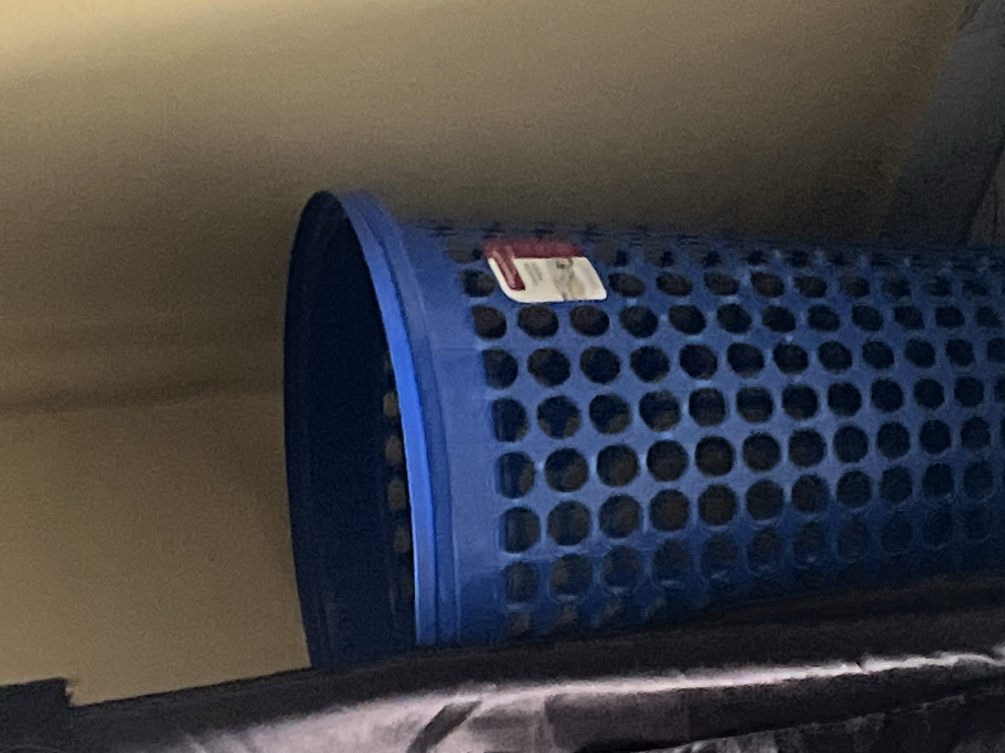 An indoor, waist-up, medium shot shows a blue plastic laundry basket with a perforated design, located next to a dark, possibly cloth-covered object, against a light brown wall. The laundry basket has a white and red label attached to its exterior. The lighting in the image is dim, casting shadows and highlighting the textures of the objects.