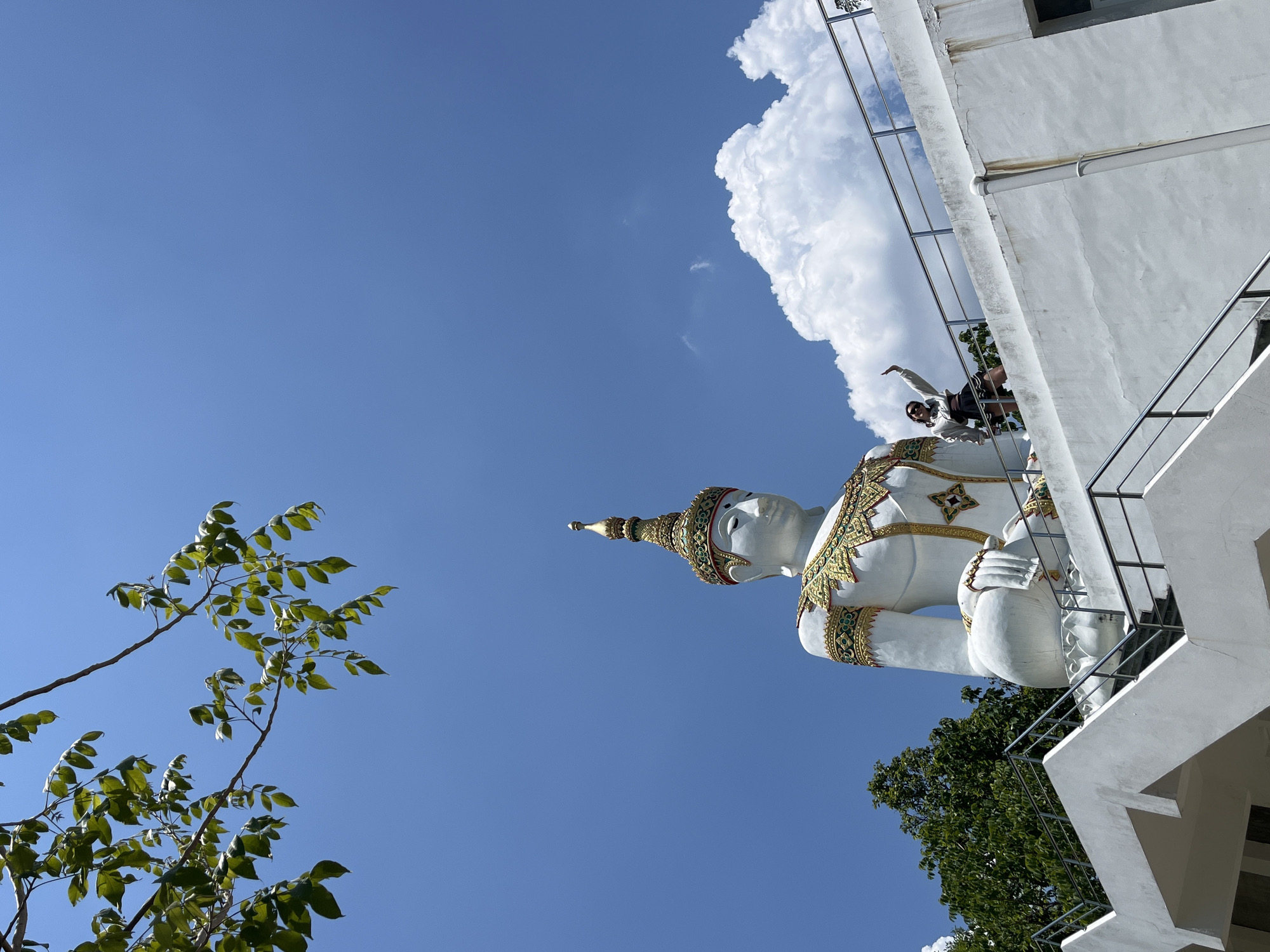 The photograph shows a large, ornate, white and gold statue in Nong Yaeng, Thailand, viewed from below. The statue is seated in a meditative pose. To the left, there are branches of a tree with green leaves. The sky is blue, with a large white cloud behind the statue. A white building with metal railings is partially visible, positioned behind and to the right of the statue. A person stands on a balcony of the building near the statue. The photo is taken from an unconventional angle, tilting upwards.
