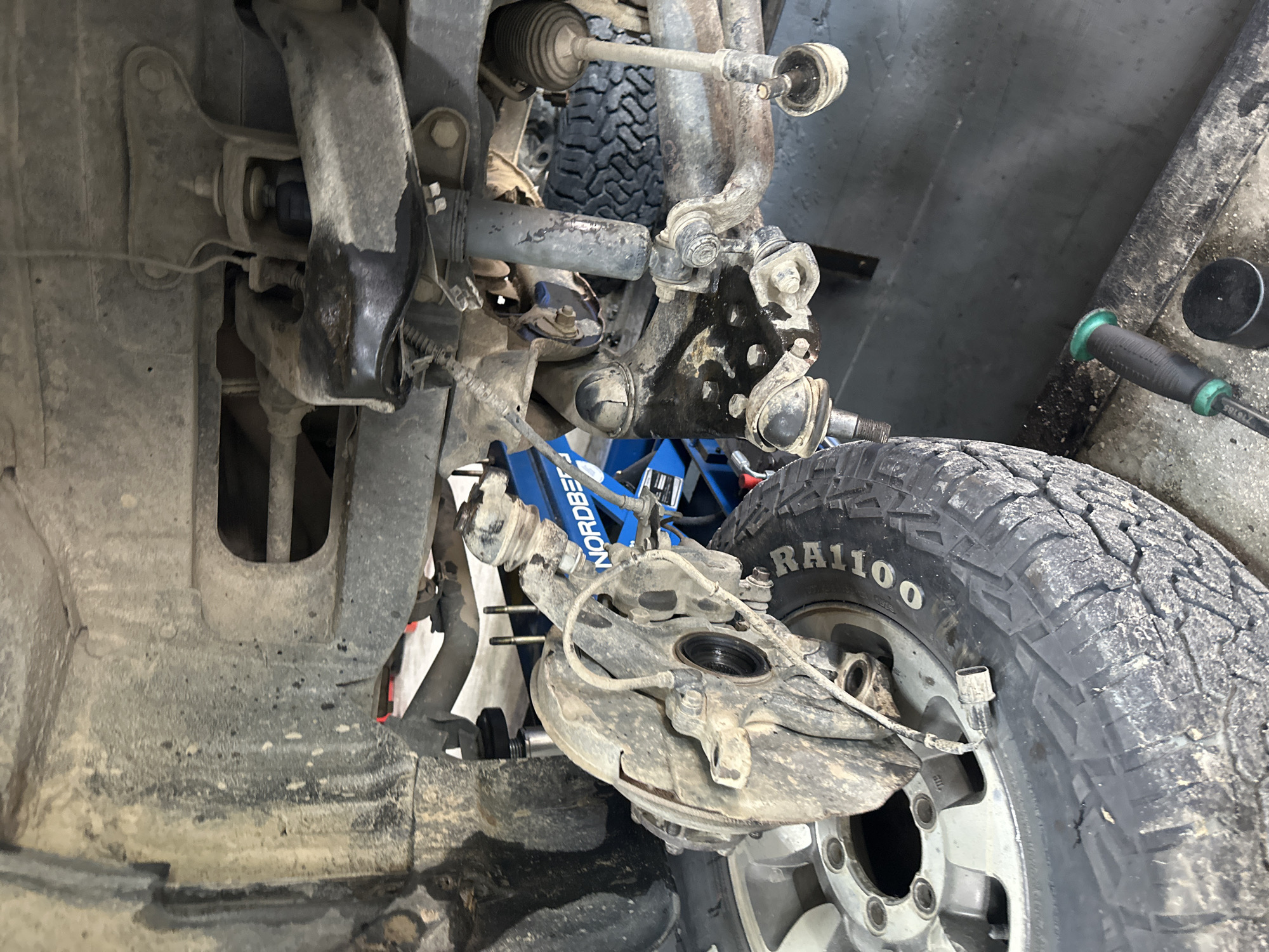 The image shows the undercarriage of a vehicle, possibly in a repair shop. A tire and wheel are visible on the left, with the brake assembly partially removed. The suspension components, including control arms, ball joints, and a shock absorber, are exposed. A blue hydraulic jack is positioned beneath the vehicle, partially obscured by suspension parts. The area appears dirty and possibly wet, with mud and grease present on the vehicle's components and the ground. Tools, including a screwdriver with a green handle and a black cylindrical object, are visible on the floor in the foreground. The background is dark and indistinct, suggesting an indoor environment. There is no visual information in the image that would indicate the location in the city of Blagoveshchensk, Russia.