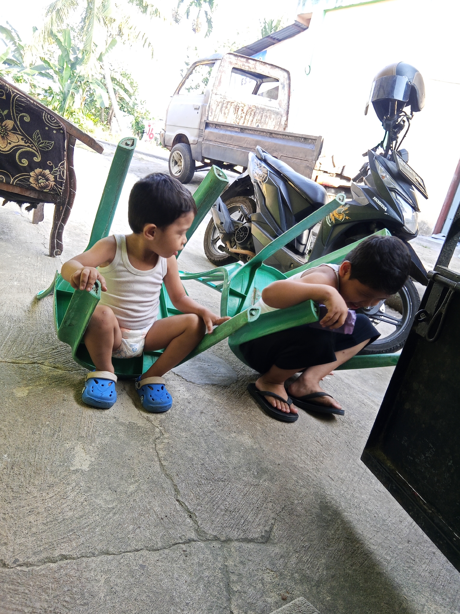 Two young boys are playing with a broken green plastic chair outdoors on a concrete surface. The boy on the left, wearing a white tank top and blue Crocs, is seated within the chair's frame. The boy on the right, wearing dark shorts and black flip-flops, is crouched beside the chair, appearing to interact with it. In the background, a dilapidated truck and a black motorcycle are visible. Lush green foliage suggests a tropical environment. The scene is likely located in the city of Lubuklinggau, Indonesia, given the presence of elements commonly found in residential areas of the region.