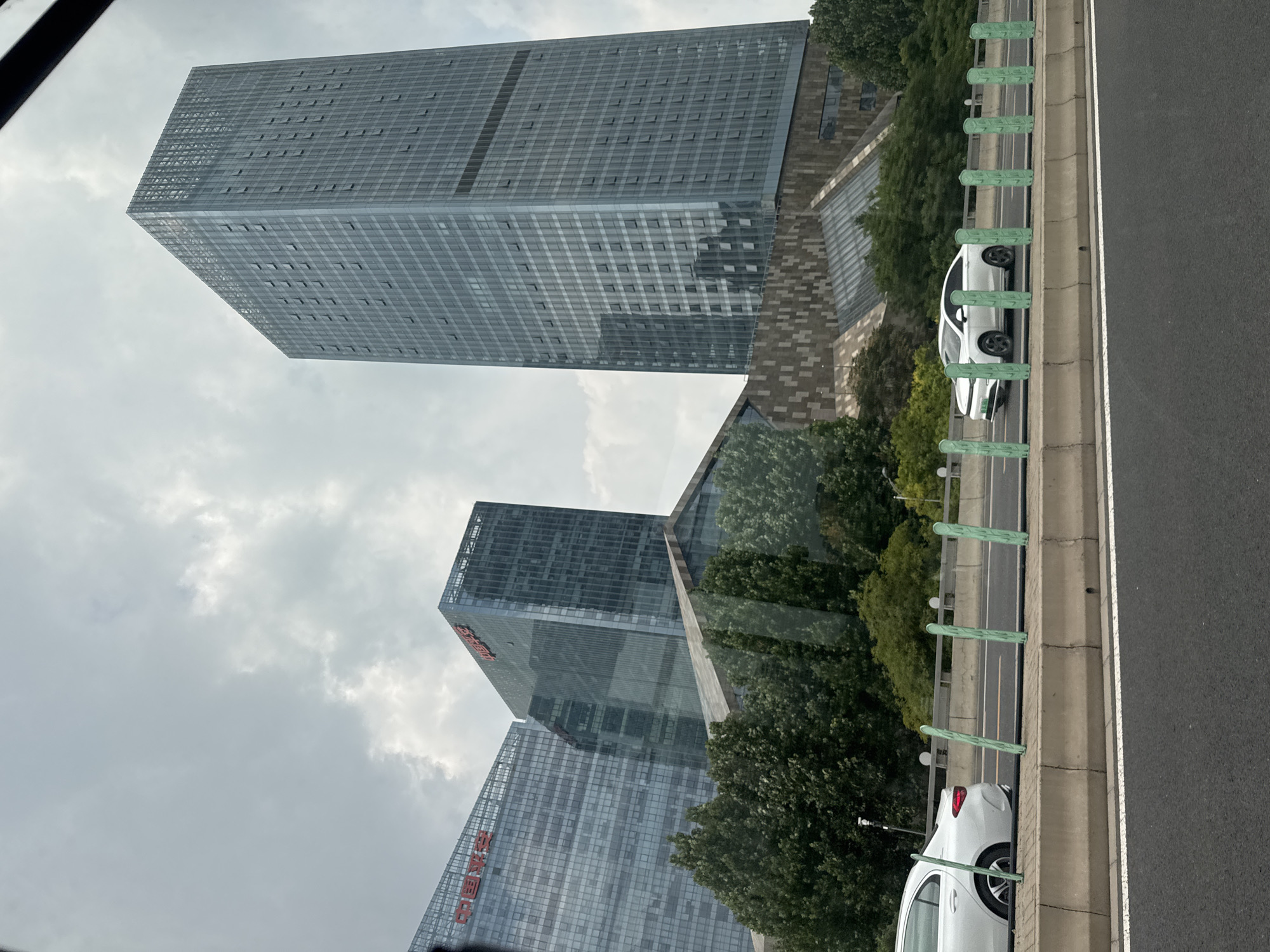 The image displays a view of modern skyscrapers and a highway in Chaoyang District, China. The sky is overcast with clouds. In the foreground, a highway with a concrete barrier and green posts is visible. Two white cars are driving on the highway. Behind the highway, a cluster of contemporary buildings, characterized by glass facades, are situated amidst lush green trees. One of the buildings, a tall, slender skyscraper, is particularly prominent on the right side of the image. Red characters, likely Chinese script, are visible on the facades of some of the buildings on the left. The overall scene suggests an urban environment with significant architectural development.