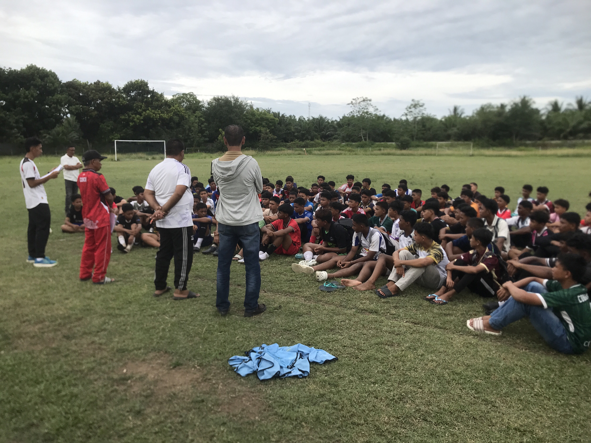 The scene shows a group of about 50 people, mostly young men, gathered on a grassy field in Tambontunong, Indonesia. They are sitting on the ground, facing a group of four adults. The adults appear to be coaching or instructing the group. The people are dressed in various clothing, including athletic wear. A soccer goal is visible in the background. The sky is overcast. A pile of blue clothing is on the ground near the center of the frame.