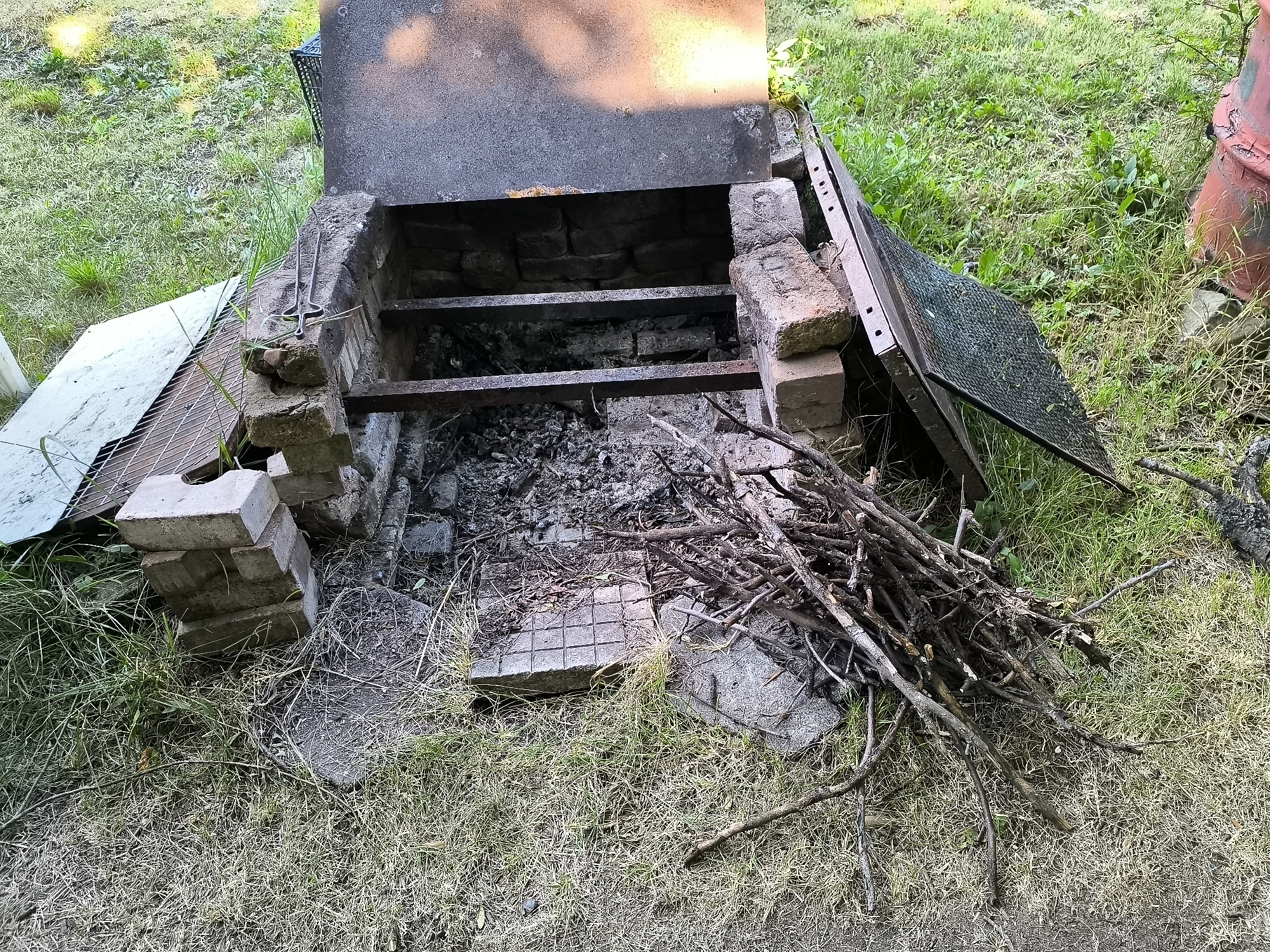 This is an overhead shot of a homemade brick barbecue grill with a metal grate and lid. The grill is located outdoors on a grassy area with some scattered debris. There is a pile of dry twigs and branches next to the grill, suggesting preparation for a fire. The grill itself appears to be old and weathered, with ash and remnants of past fires visible inside. The surrounding environment is a lawn with patches of dry and green grass. The scene is located in Sofia, Bulgaria.