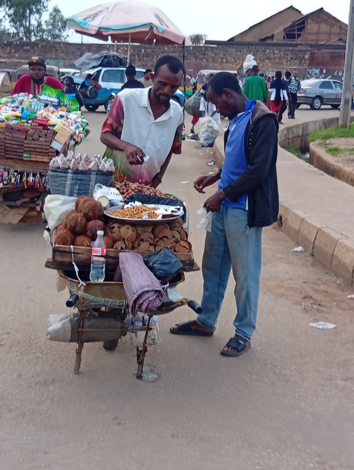 In Bukuru, Nigeria, two men are interacting next to a street vendor's cart. The man on the left, wearing a white shirt and a colorful patterned smock, is attending to the cart's wares. The cart itself is laden with coconuts, a dish of nuts, and various packaged goods. To the right, a man in a blue shirt and black jacket is facing the vendor, appearing to engage in a transaction or conversation. He is holding a plastic bag. The background reveals a street scene with other pedestrians, vehicles including a car and a mini-van, and buildings. The ground is a dusty road surface, and a concrete curb runs alongside the pavement. The atmosphere suggests a bustling marketplace or roadside commercial area.