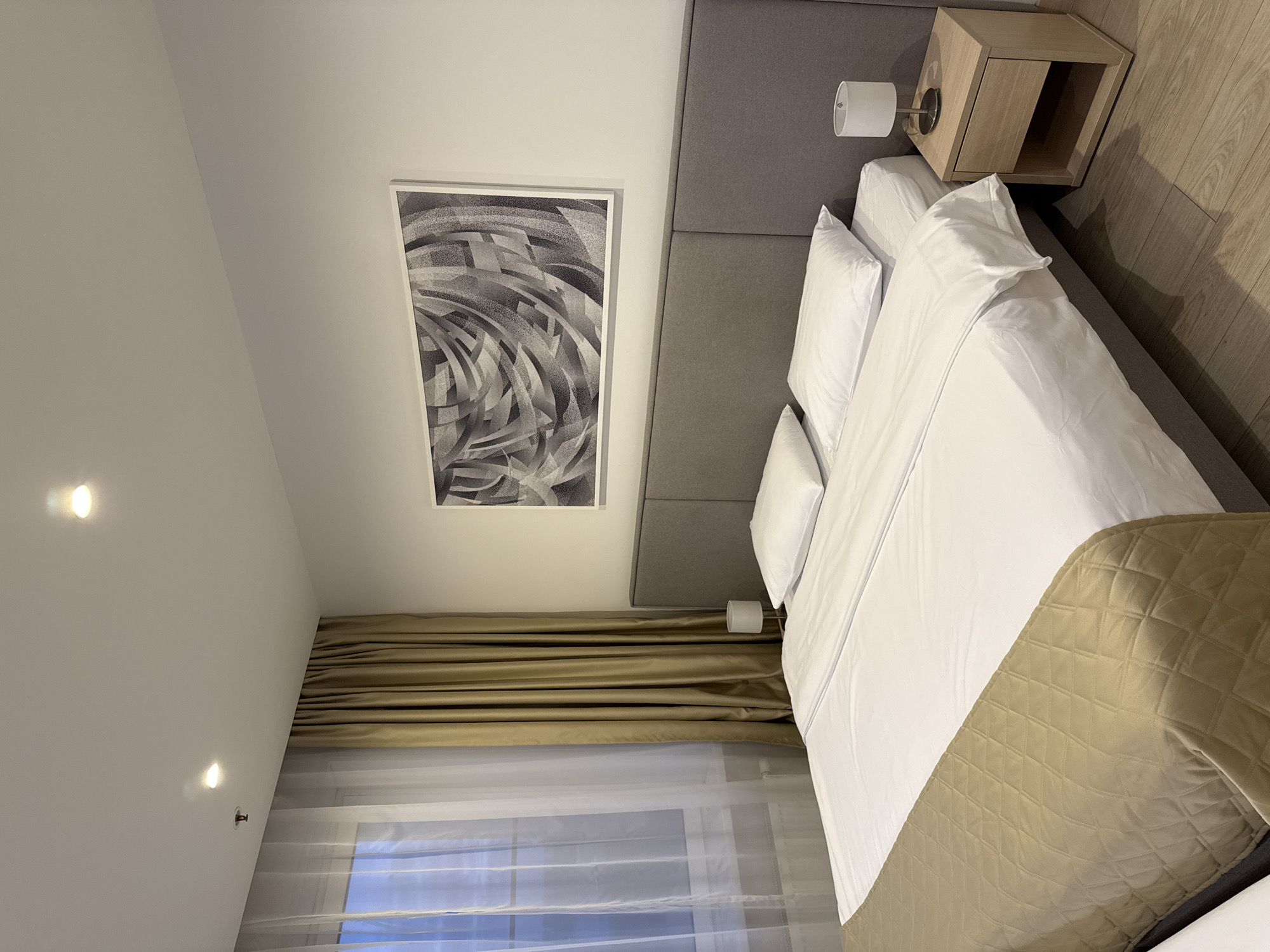 A bedroom is shown from an elevated perspective. The primary focus is a bed with white linens and a gold quilted bed runner. The bed is positioned against a gray headboard, with a light-colored nightstand on the right side of the bed. Above the headboard is an abstract black and white artwork on the wall.
Curtains with a combination of sheer and solid gold fabric cover the window behind the bed. The walls are a light neutral color, and recessed lighting is visible in the ceiling. The floor appears to be light-colored wood.
The location is not discernible from the given information.