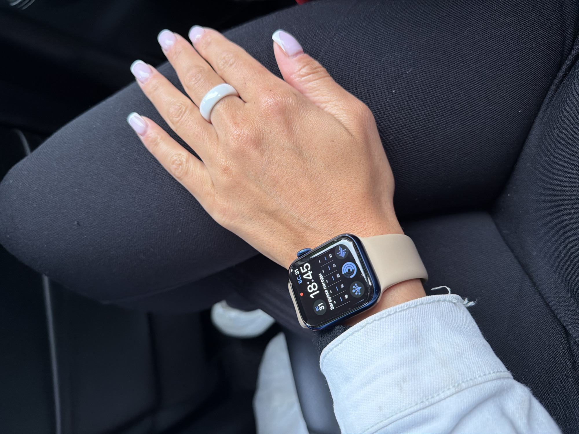 A woman's left hand and wrist are prominently displayed against a background of dark, possibly black, fabric. She is wearing a white ceramic ring on her ring finger, and her nails have a short, square shape with a white tip. The hand rests on the leg, which is clad in dark, fitted trousers or leggings. On her wrist, she sports a blue Apple Watch with a tan-colored band. The watch display shows a digital clock reading "18:45" and other informational icons. A small portion of a white denim jacket sleeve peeks out from under the watch. The setting appears to be indoors, likely a car interior, as suggested by the seat fabric and partial view of a white object in the lower center. There is no information in the image about location in the city of Pushkino, Russia.