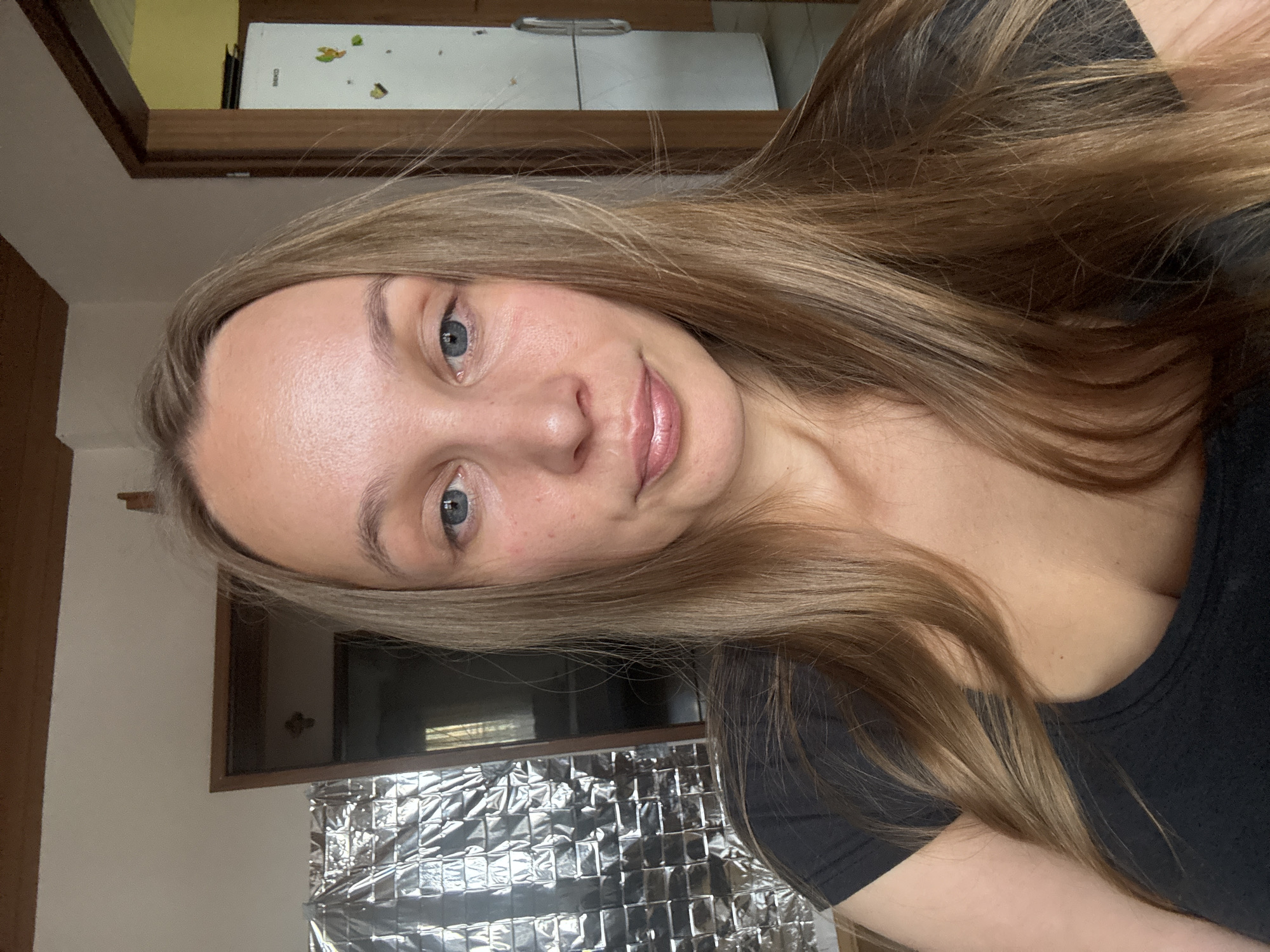 A medium shot, taken from a slightly high angle, captures a Caucasian woman with long, straight, honey-blonde hair and blue eyes. She is wearing a dark grey t-shirt with a wide neckline. Her skin has a fair complexion with some subtle redness and a few visible pores. Her lips are slightly parted and appear to be glossed.
The background suggests an indoor setting, possibly a home. To the left of the woman, a large, metallic, crinkled material is visible, possibly insulation or a reflective surface. Behind her, a doorway leads to another room, and to the right, a white refrigerator with a silver handle stands against a wall. The overall lighting is natural and diffused. There are no specific landmarks indicating a particular location in Überherrn, Germany, as the scene appears to be a generic indoor environment.