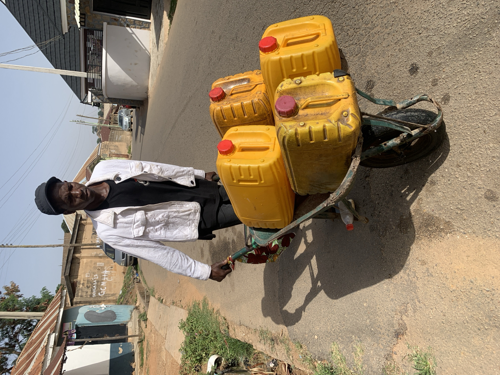 A man stands on a street in Jos, Nigeria, pulling a wheelbarrow loaded with four yellow jerrycans with red caps. The man wears a black shirt, white jacket, and black cap. The wheelbarrow is old and weathered, with a green frame and a worn tire. The street is paved but shows signs of wear and tear. Buildings and vegetation line the sides of the street.