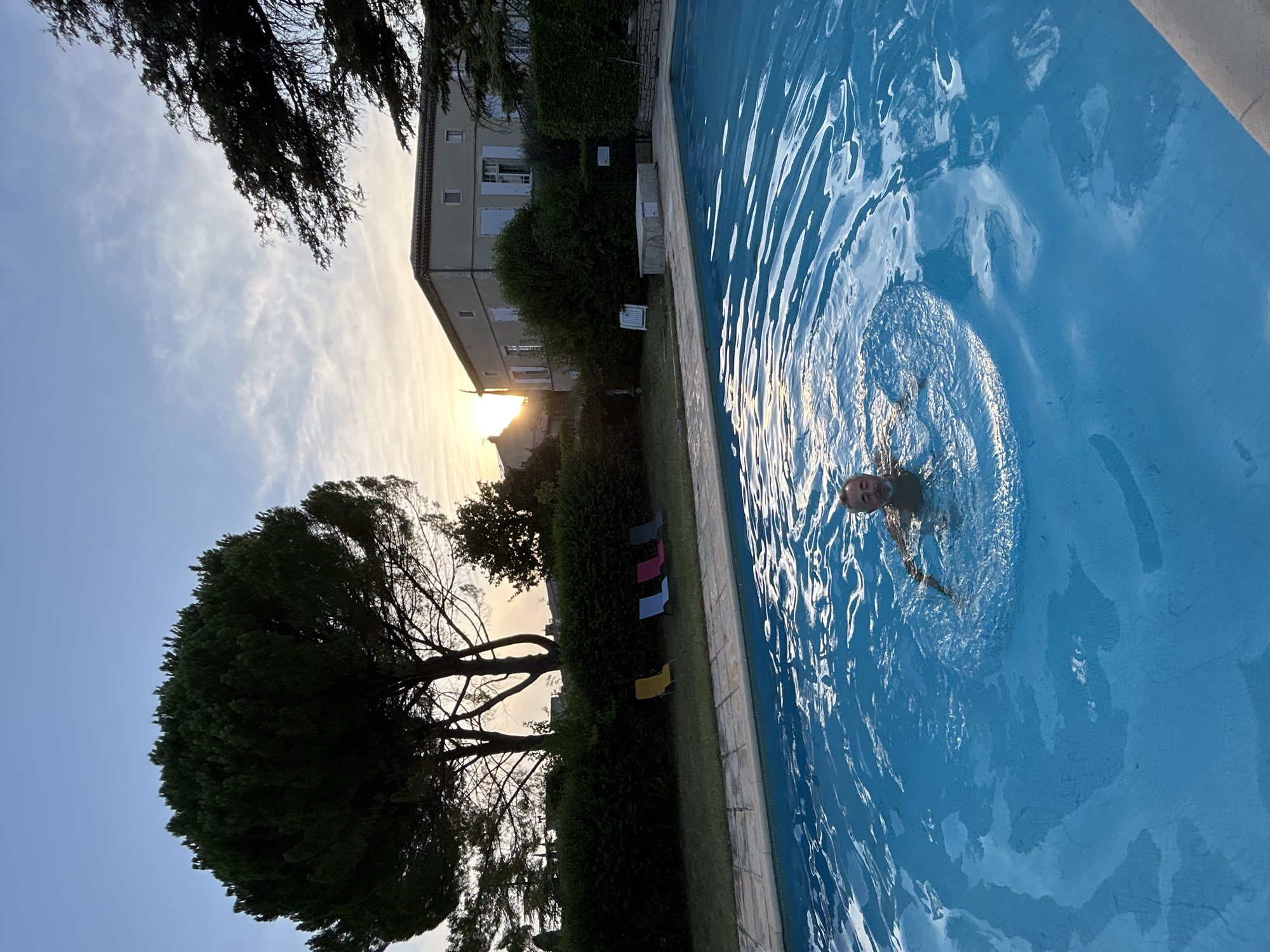 The image depicts a swimming pool with a man swimming in it, located in Saint-Marcel-lès-Sauzet, France. The sun is setting, casting a warm glow on the scene. In the background, a building with a tiled roof and light-colored walls is visible, surrounded by trees and hedges. There are several lounge chairs arranged on the grass next to the pool. The water in the pool is a vibrant blue, with ripples indicating movement. The man, partially submerged in the water, is facing the camera and appears to be smiling. The overall atmosphere is serene and relaxed, suggesting a vacation or a peaceful evening.