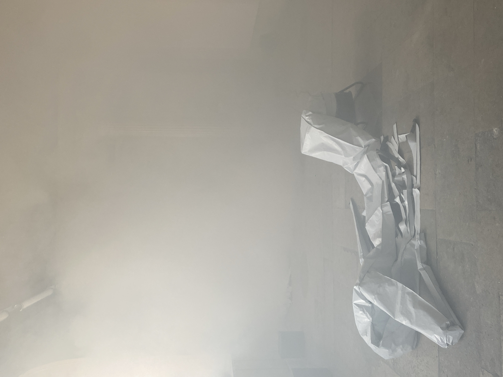 The image shows a room filled with smoke. On the right side of the frame, a white, crumpled object, potentially a garment or protective suit, lies on the floor. The floor appears to be tiled. In the background, on the left, a pipe is visible through the smoke. The overall visibility is low due to the heavy smoke. It is impossible to ascertain the location of the scene in Paris, France.