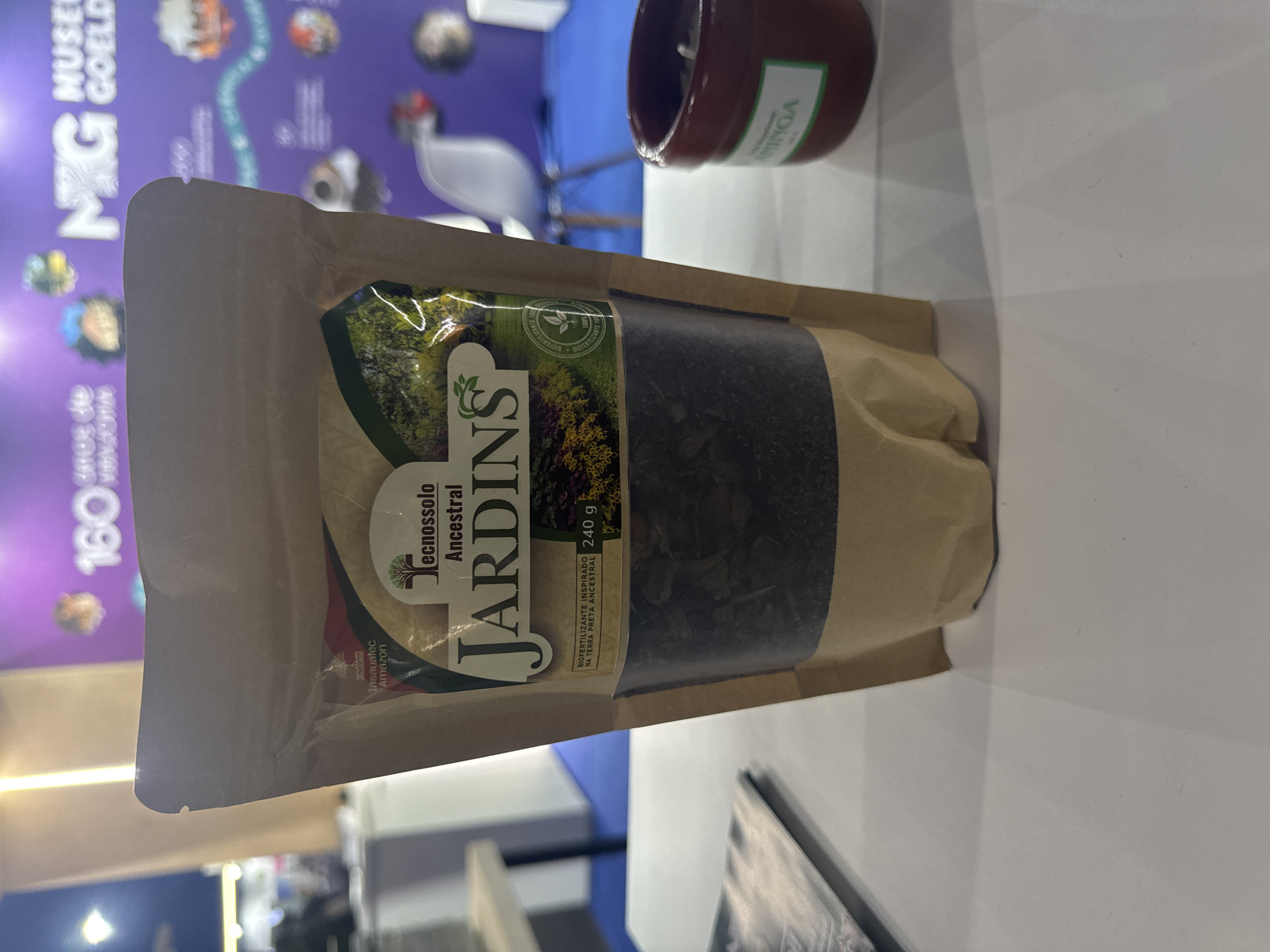 The image shows a close-up of a brown paper bag containing what appears to be a dark, granular product. The bag has a label that reads "Tecnossolo Ancestral Jardins" in a stylized font, with images of plants and soil. Below this, smaller text indicates "BIOFERTILIZANTE INSPIRADO NA TERRA PRETA ANCESTRAL" and "240 g". The bag is sealed at the top. In the background, a blurred purple wall features text and graphics that appear to be from an exhibition or museum, possibly related to "MUSEU GOELDI" and a "160 anos de vida". To the right of the bag, a small, reddish-brown ceramic pot with a white label is visible. The scene appears to be indoors, possibly at an exhibition booth or a store display. There are no visible people or identifiable landmarks of Niterói, Brazil.