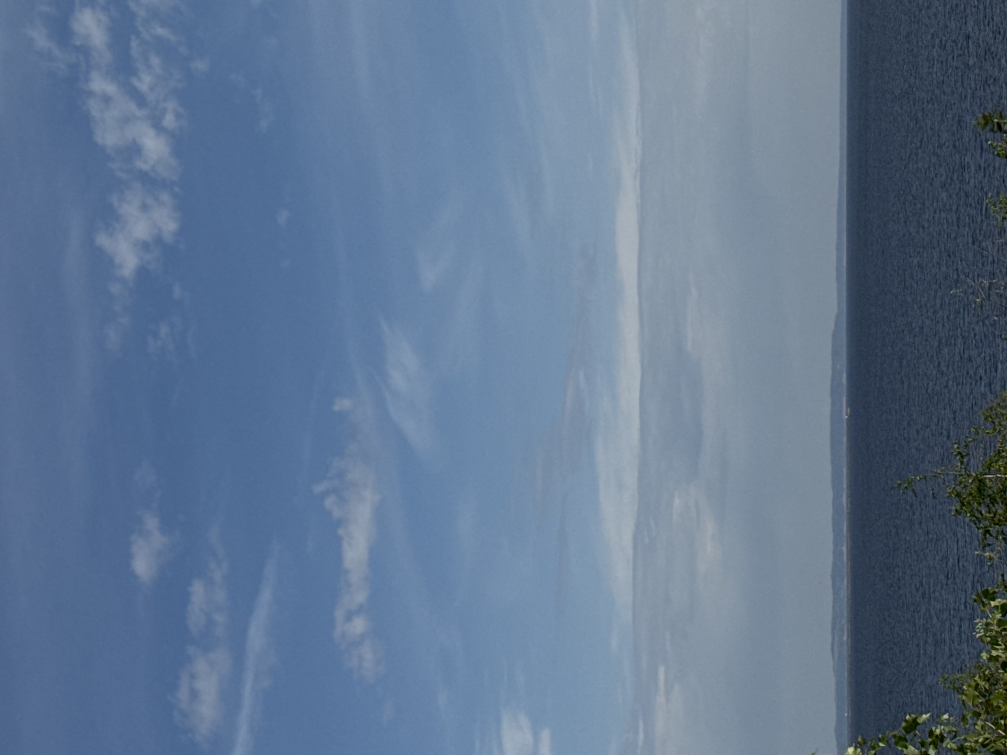 The image shows a view of the Kama River in Chaikovsky, Russia. The sky is predominantly blue with scattered white clouds. The river stretches along the right side of the image, with a darker blue color. Some greenery is visible on the right edge of the frame, possibly from trees or bushes along the shoreline. The image appears to be taken on a clear, sunny day.