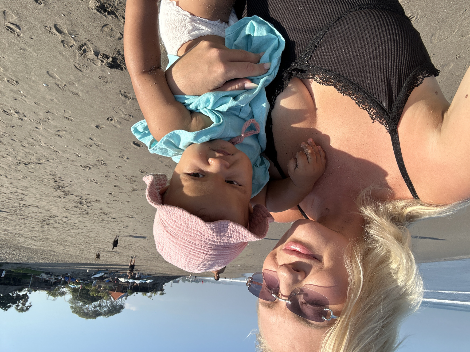 A blonde woman wearing sunglasses and a black tank top holds a baby on a beach in North Kuta, Indonesia. The woman is smiling, and the baby is wearing a pink hat and a blue outfit. The beach is sandy, with footprints visible. In the background, there are trees, buildings, and people walking. The ocean is visible in the distance. Two dogs are running on the beach.