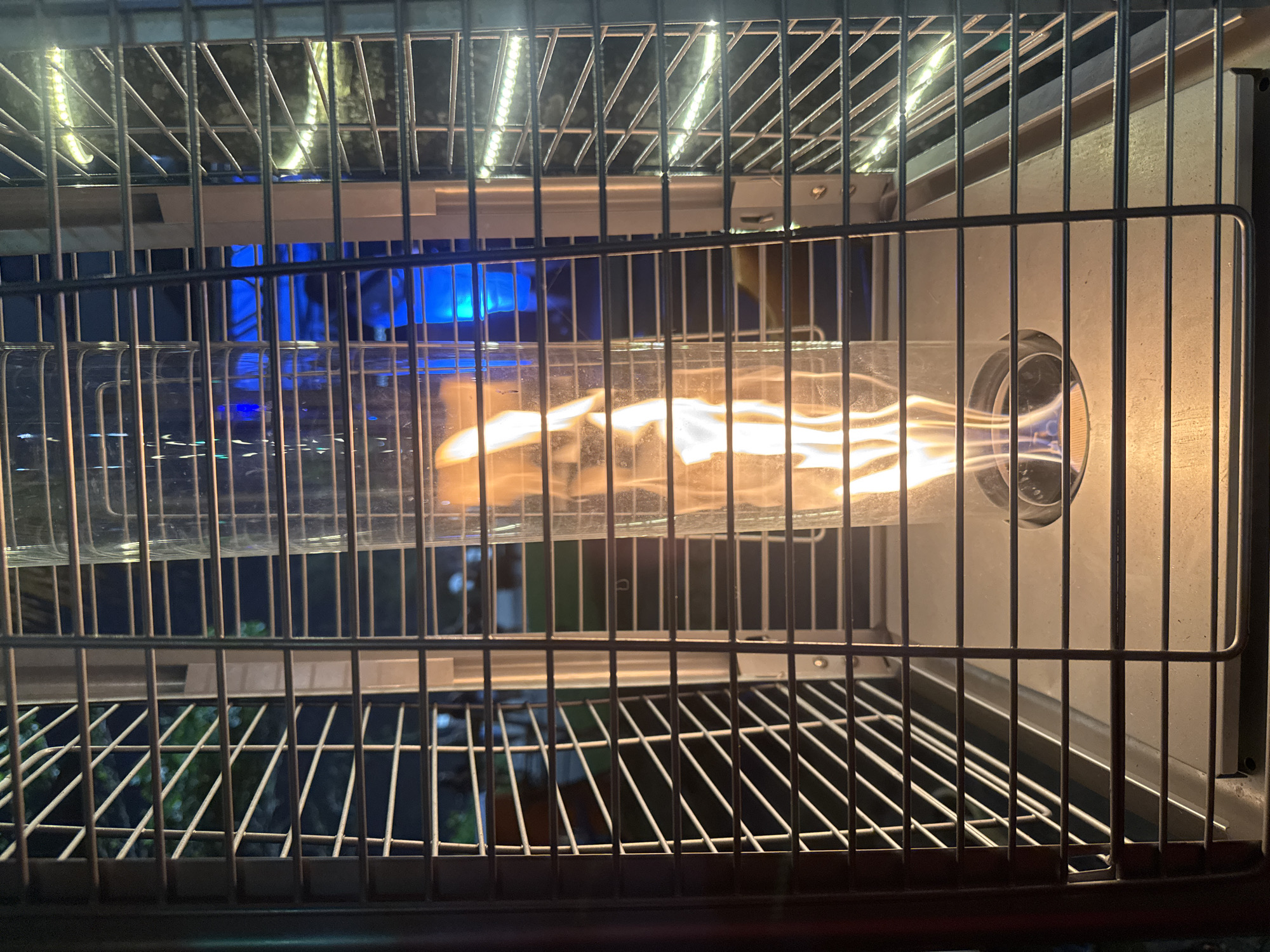 The image shows a close-up view of a propane patio heater with a visible flame inside a glass cylinder. The heater is enclosed by a metal grate. In the background, there is blue lighting, possibly from a bar or restaurant, and some greenery. The overall scene suggests an outdoor dining or entertainment area at night. The location is an outdoor restaurant in Niterói, Brazil.