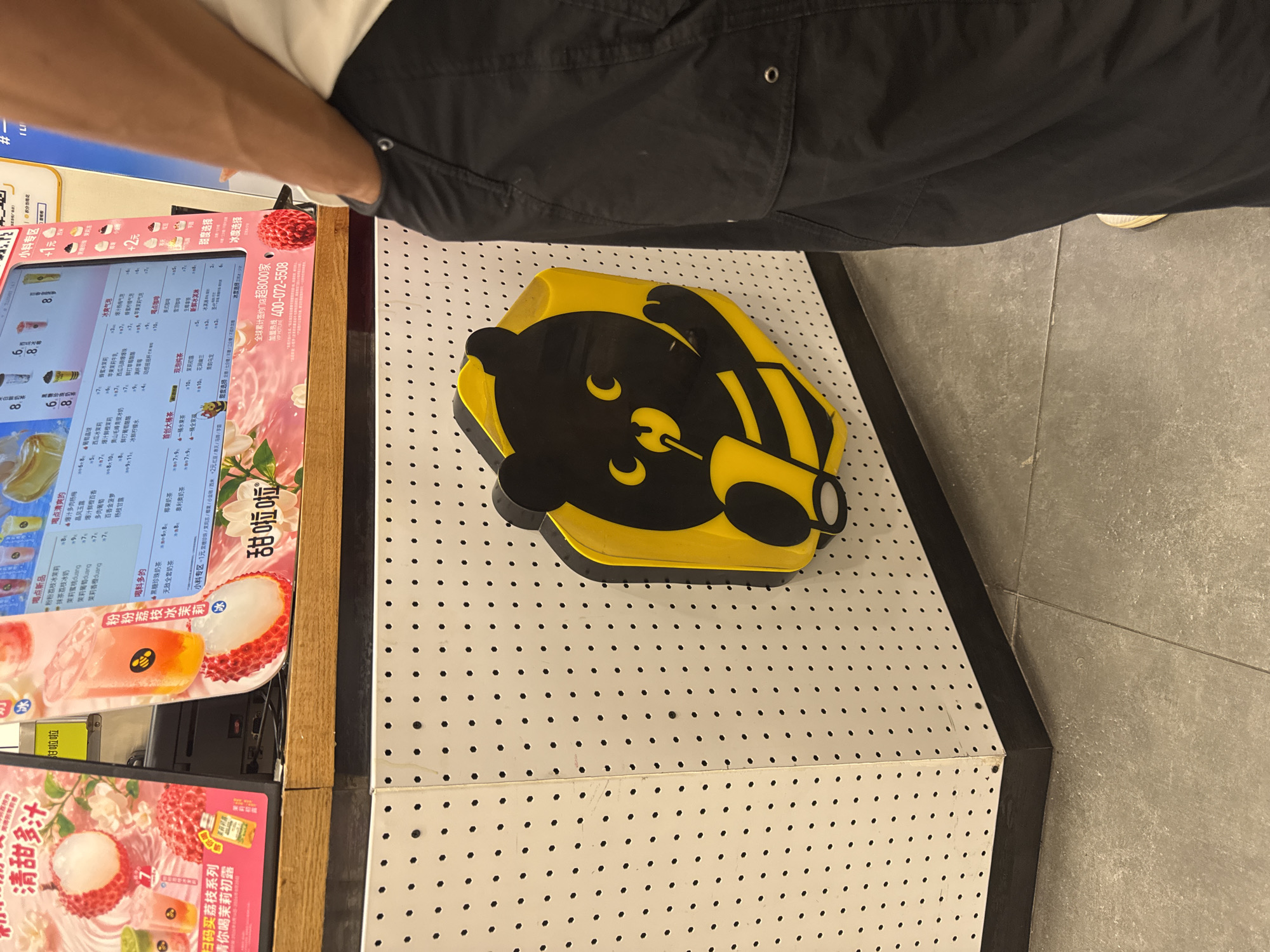 The image shows a close-up view of a beverage shop counter in Shenyang City, China. A black and yellow hexagonal sign featuring a cartoon bear drinking from a yellow cup is mounted on a white pegboard wall. The counter also displays promotional posters for various drinks, including lychee-themed beverages. A person wearing black pants is standing to the right of the sign, partially obscuring the counter. The floor is tiled with a grey, stone-like pattern.