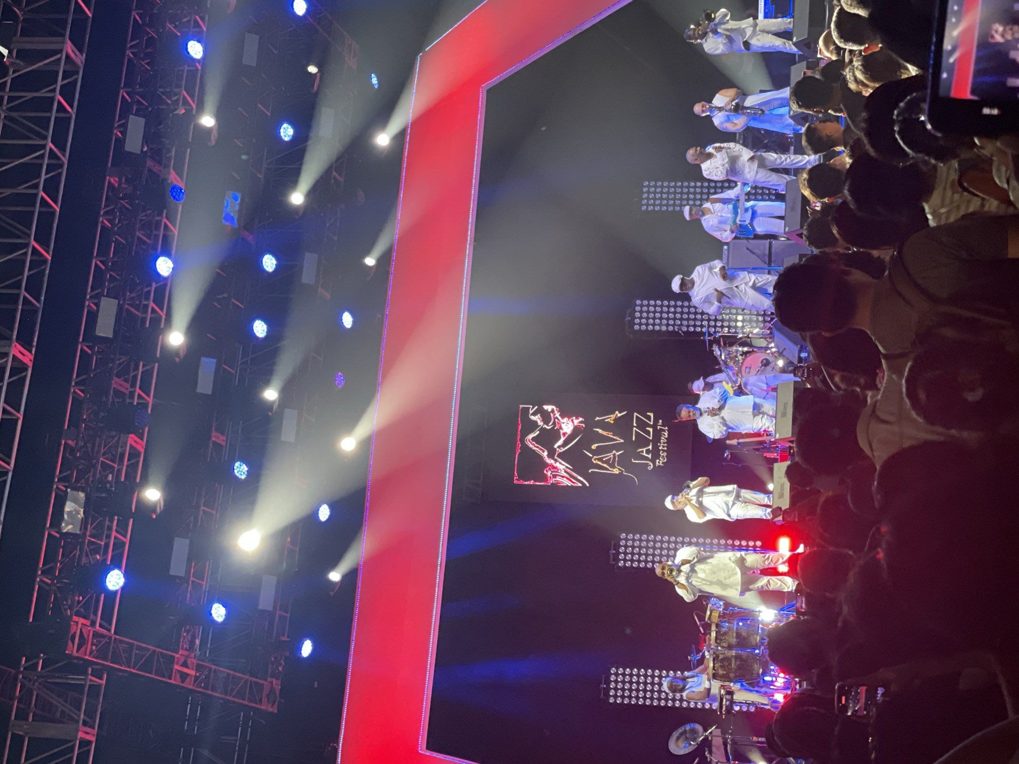 A concert is being held in Salembaran, Indonesia, with a live jazz band performing on stage in front of a crowd. The band members are dressed in white and playing various instruments, including trumpets, saxophones, guitars, and drums. The stage is lit with bright white and blue lights, and there's a large screen displaying the Java Jazz Festival logo behind the band. There is also a red vertical block on the left of the image. The crowd is dense, with people holding up their phones to record the performance. The overall atmosphere is vibrant and festive.