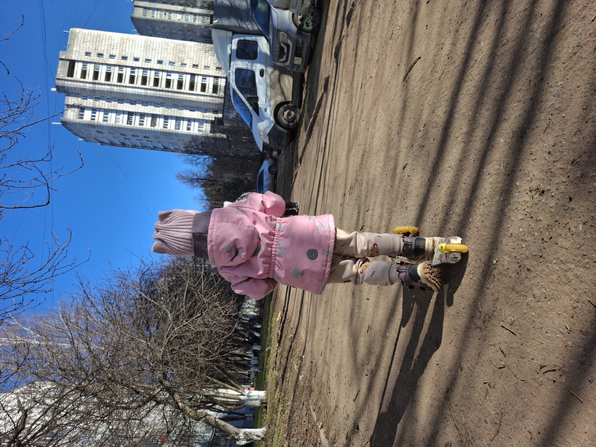 A small child stands on roller skates on a dirt path. The child wears a pink jacket, light-colored pants, and a pink knit hat. A tall apartment building is visible in the background, along with a white van. Bare trees are on the left side of the frame. The location appears to be in the city of Chaikovsky, Russia.