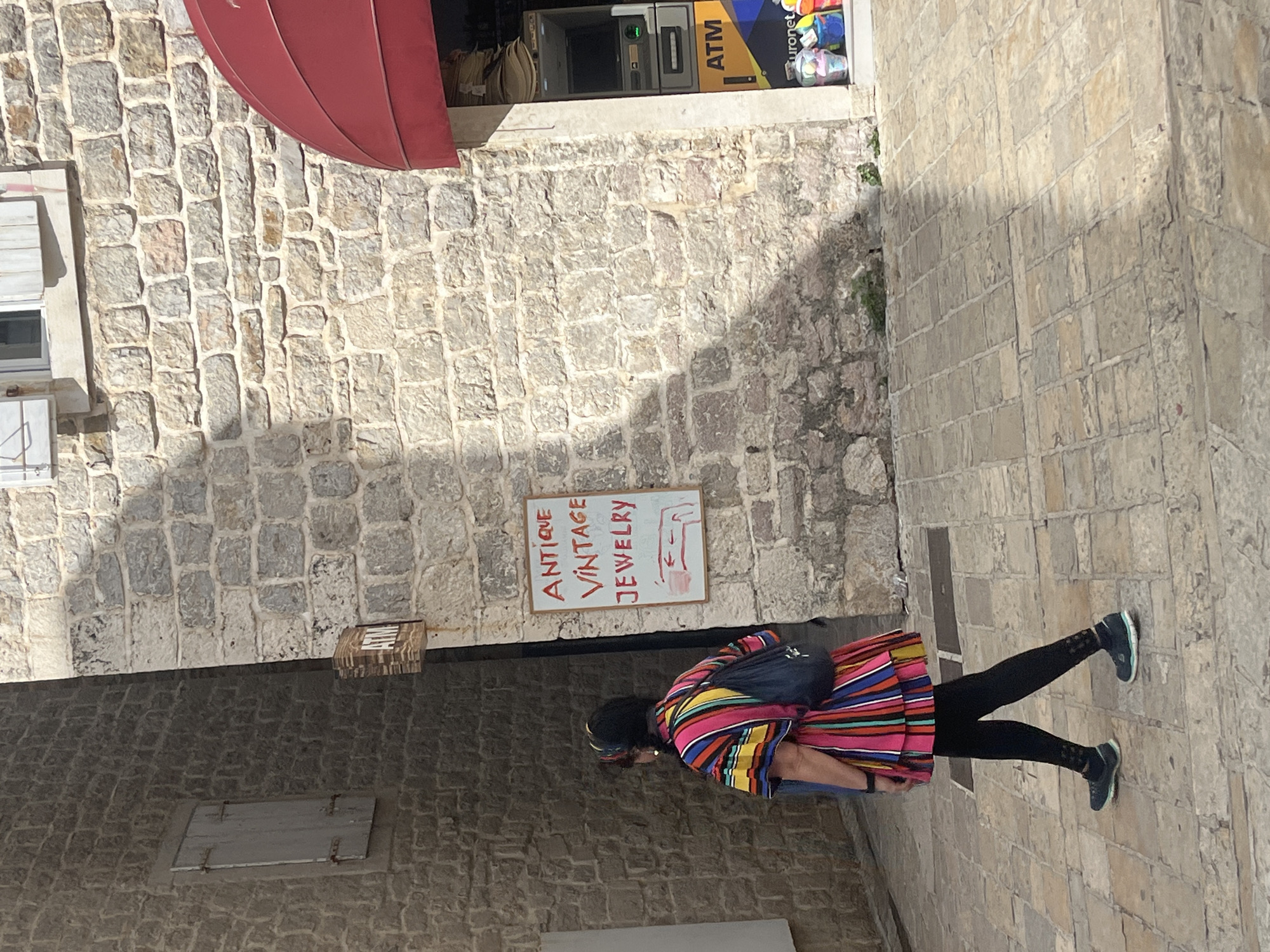 This image captures a bright, sunlit narrow street in what appears to be the historic area of Budva, Montenegro. The street is paved with light-colored cobblestones and flanked by sturdy stone buildings with irregular, grey and beige blocks.
In the foreground, a woman walks away from the viewer, slightly off-center to the right. She is wearing black leggings, dark athletic shoes with light soles, and a distinctive, brightly colored poncho-style top featuring horizontal stripes of pink, blue, yellow, orange, and green. She carries a dark blue backpack.
The architecture is traditional, with a building on the left featuring a small white window with closed wooden shutters. A red fabric awning extends over an entrance higher up on the left side of the frame. Further down the street, an ATM is visible inside a shop window, identified by the prominent yellow "ATM" text and the "Euronet" logo above it.
On the stone wall to the left of the woman, a white rectangular sign with red lettering clearly states "ANTIQUE VINTAGE JEWELRY." Below the text, a simple red drawing of a hand with a pointed finger indicates direction. The sunlight casts distinct shadows on the ground and walls, highlighting the texture of the stone. The scene suggests a pleasant daytime atmosphere in a historical setting.