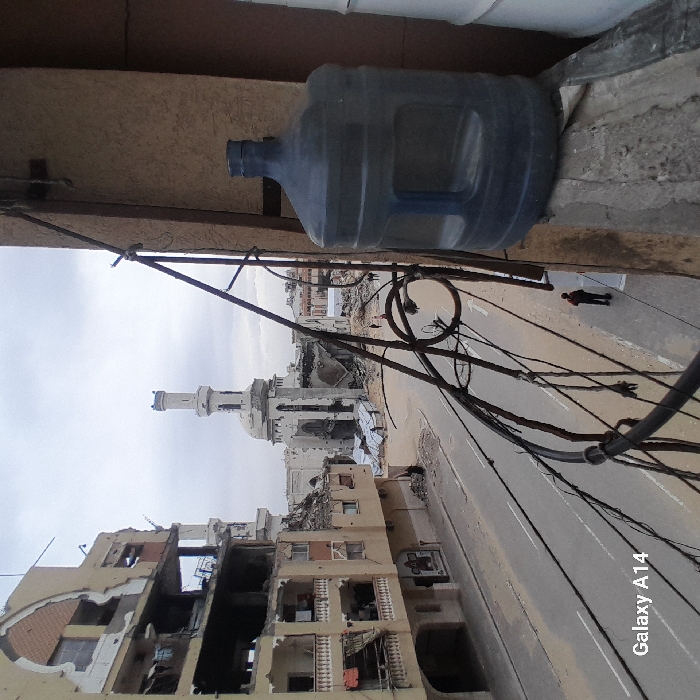 The image captures a scene of extensive urban destruction in Khan Yunis, Palestinian Territory, viewed from an elevated perspective, likely a balcony. In the immediate foreground on the right, a large, translucent blue 5-gallon plastic water bottle rests on a concrete ledge. A dark metal railing with multiple intertwined black cables extends diagonally across the right side of the frame.
Across the street, a multi-story light-brown residential or commercial building on the left exhibits severe structural damage, with several floors exposed due to missing exterior walls and damaged balconies. Rubble is visible at its base. Dominating the background is a white mosque with a tall minaret, also extensively damaged; its dome appears partially collapsed, and large sections of its walls are missing, revealing the interior. Piles of debris surround the mosque.
The asphalt street below is largely clear of traffic, though scattered debris lines its edges. A white arrow indicating direction is painted on the road. One individual in dark clothing stands on the sidewalk in the lower mid-ground, facing away from the camera. Additional small, indistinct figures are visible near the mosque further down the street. The sky is uniformly overcast and grey.