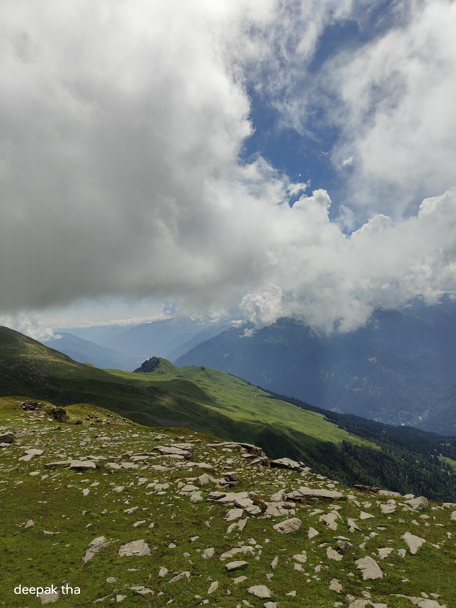 A wide-angle, full shot captures a mountainous landscape in an overcast, daytime setting. The foreground is dominated by a grassy hill dotted with scattered rocks and stones. The midground features rolling green hills that recede into the distance, with shadows cast across the slopes. Further back, multiple layers of mountains are visible, fading into a hazy blue. The sky is a mix of bright blue patches and heavy, grey clouds, with some sunlight breaking through in the distance. The image has a vertical orientation. The location is identified as Bashisht, India.