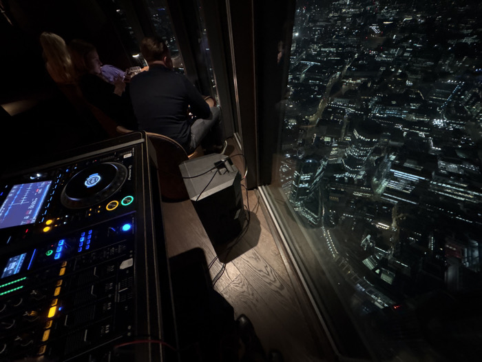 The image displays an interior scene at night from a high-rise establishment, overlooking the illuminated City of London, United Kingdom. In the left foreground, a professional DJ setup, specifically a Pioneer DJ controller with active illuminated screens and controls, is positioned on a dark surface. To the right of the DJ equipment, a compact, dark monitor speaker rests on a polished wooden floor, connected by visible cables. Further right, a male subject is seated on a stool, viewed from behind, wearing a dark long-sleeved shirt and trousers. He holds a clear glass with a beverage in his right hand and is oriented towards a large window. Partially obscured by the male subject and to his left, a second individual, likely female with blonde hair, is also visible, holding a glass and facing the window. The background, visible through the expansive window, presents a panoramic night view of London's urban landscape, densely populated with illuminated buildings, streets, and vehicle lights, consistent with a high-level perspective from a venue within The Shard.