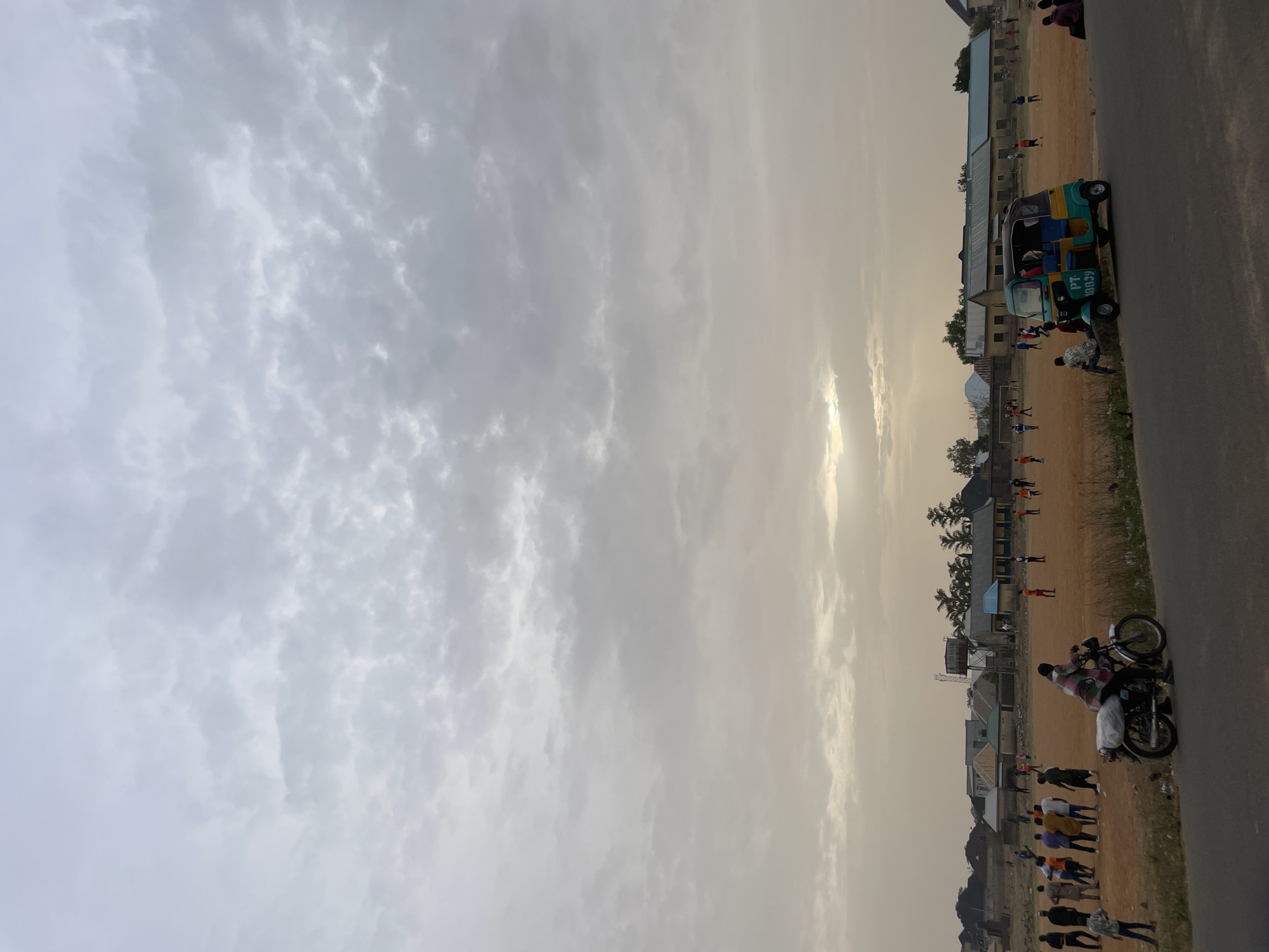 The image captures a scene in Jos, Nigeria, under a heavily clouded sky. A dirt field stretches horizontally, with numerous individuals scattered across it, possibly engaged in recreational activities. Adjacent to the field is a paved road. A distinct blue and yellow auto-rickshaw is parked alongside the road. A motorcycle and its rider are on the lower right portion. Structures, likely buildings, are present along the edge of the field. The lighting suggests either dawn or dusk.