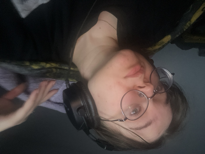 An indoor, waist-up, angled portrait features one young adult female with short, dark hair and round-frame glasses. She wears a dark t-shirt or top and a thin, light-colored necklace. Black over-ear headphones are positioned on her head, with a cable visible descending from the right earcup. Her head is tilted slightly to the viewer's left, and her gaze is directed upwards and to the viewer's right. Her expression is neutral. The subject's right hand is visible in the upper right portion of the image, resting on a purple or lavender-colored fabric. Below this, and to the subject's left, a dark fabric with a yellow or green pattern is partially visible. The background is dark and indistinct, suggesting low ambient light, with the subject's face illuminated. The specific location within Pushchino, Russia, cannot be determined from the visual information provided.