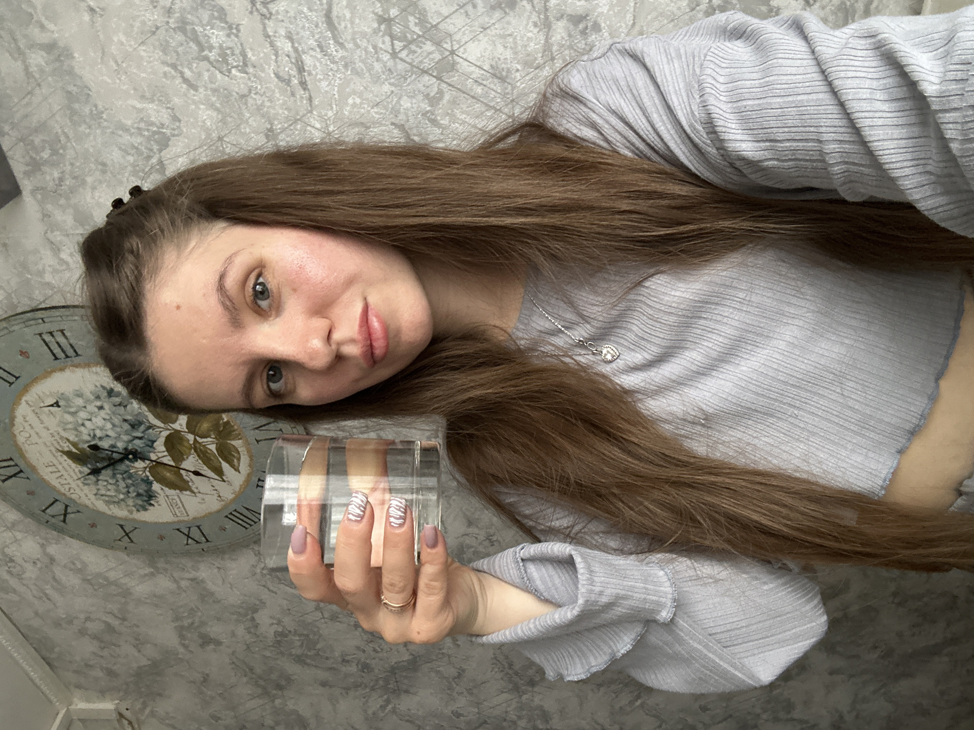 A woman with long, light brown hair, wearing a light blue ribbed shirt and a silver necklace, is holding a rectangular glass object with horizontal beige and brown stripes in her right hand. She has light-colored nails, some painted a solid light purple and others with a white and gray zebra print. She is wearing a gold ring on her ring finger. Behind her, a wall features a grey and white patterned wallpaper. A large decorative clock with Roman numerals and a floral design is visible on the wall to her left. Based on prior information, this scene could be in Klin, Russia.