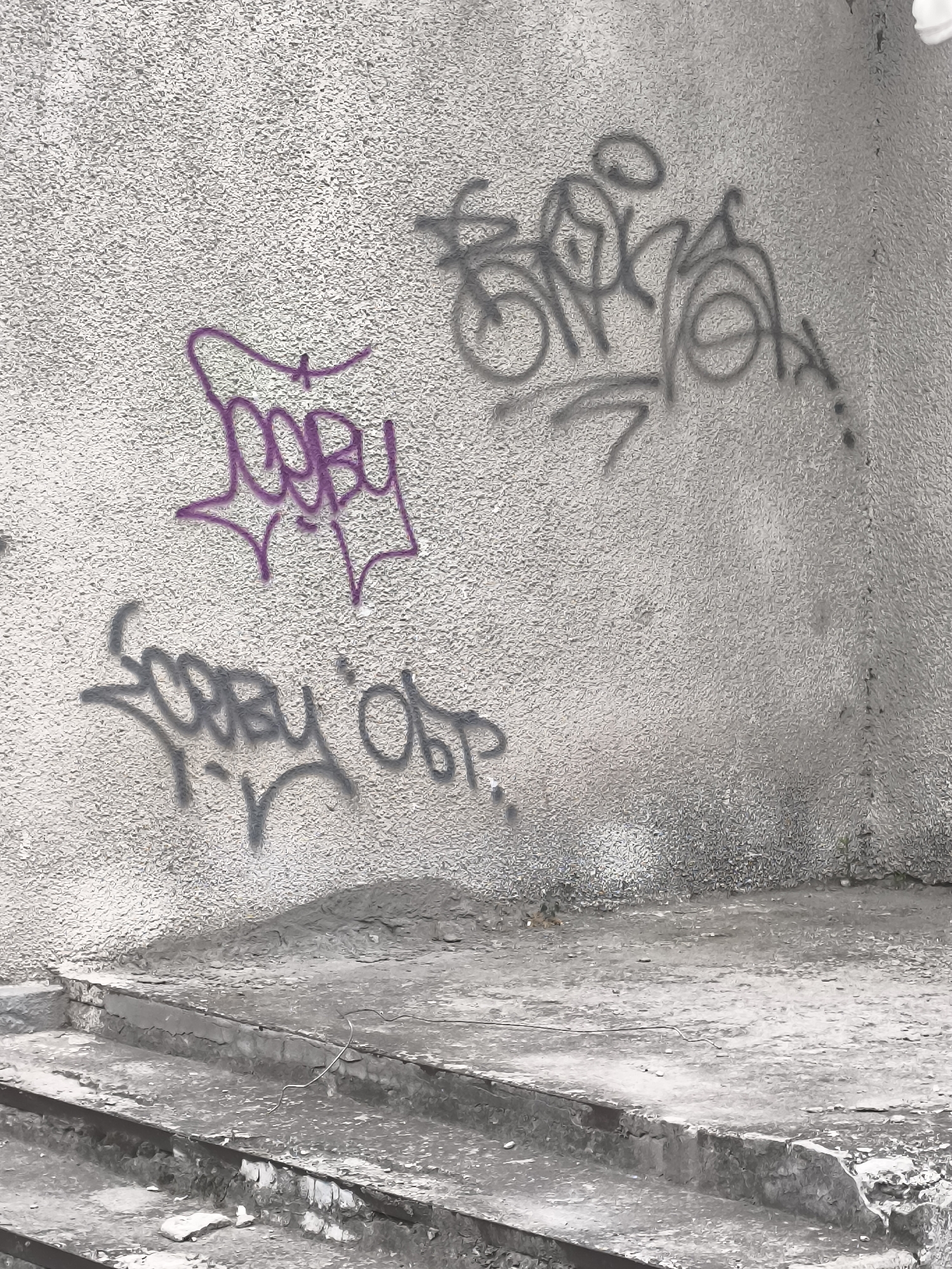 The image shows an outdoor scene with a textured, light-colored wall on the right and a rough concrete staircase in the foreground. The wall is adorned with graffiti. Two distinct graffiti tags are visible: one in purple, characterized by bubble-like lettering with sharp points extending outward, and another in a darker gray, which appears to be a more complex, stylized script with looping lines and curves. The concrete steps show signs of wear and tear, with some areas appearing chipped and weathered. The overall environment suggests an urban setting, likely a public space or an abandoned area within the city of Bălți, Moldova. There are no people or animals visible in the frame.