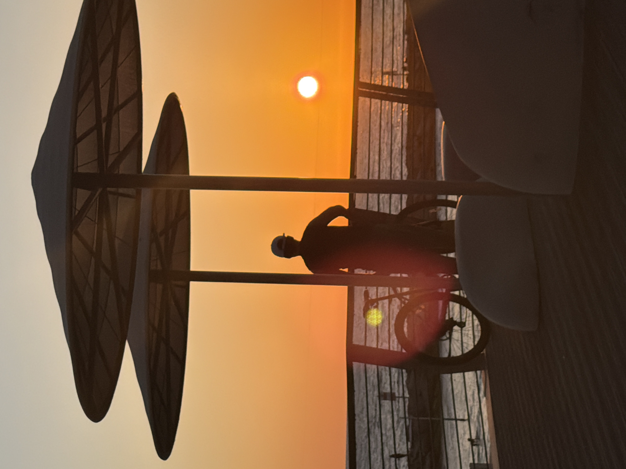 The image captures a vibrant sunset or sunrise scene over the coast of Tel-Aviv, Israel, rendered primarily in silhouette against a warm-toned sky. The sun, appearing as a bright white disk with a subtle red aura, is prominently positioned in the upper right of the frame, illuminating the sky with intense orange and yellow hues.
In the upper left, two large, dark beach umbrellas or similar shade structures are visible, their skeletal frames discernible through the fabric. A horizontal bar stretches across the mid-section of the image, forming part of the foreground architecture.
Centered in the midground, a lone individual on a bicycle is captured in stark silhouette. The person appears to be wearing a helmet or cap and is positioned as if riding along a path or boardwalk. Their form and the bicycle are sharply defined against the brilliant sky.
On the right side and extending towards the bottom, a dark, vertically slatted structure, possibly a fence, railing, or part of a building, is visible. Beyond this structure, a body of water, likely the sea or ocean, reflects some of the sky's light but remains largely muted in tone. The scene evokes a serene coastal moment at dusk or dawn. No text is visible in the image.