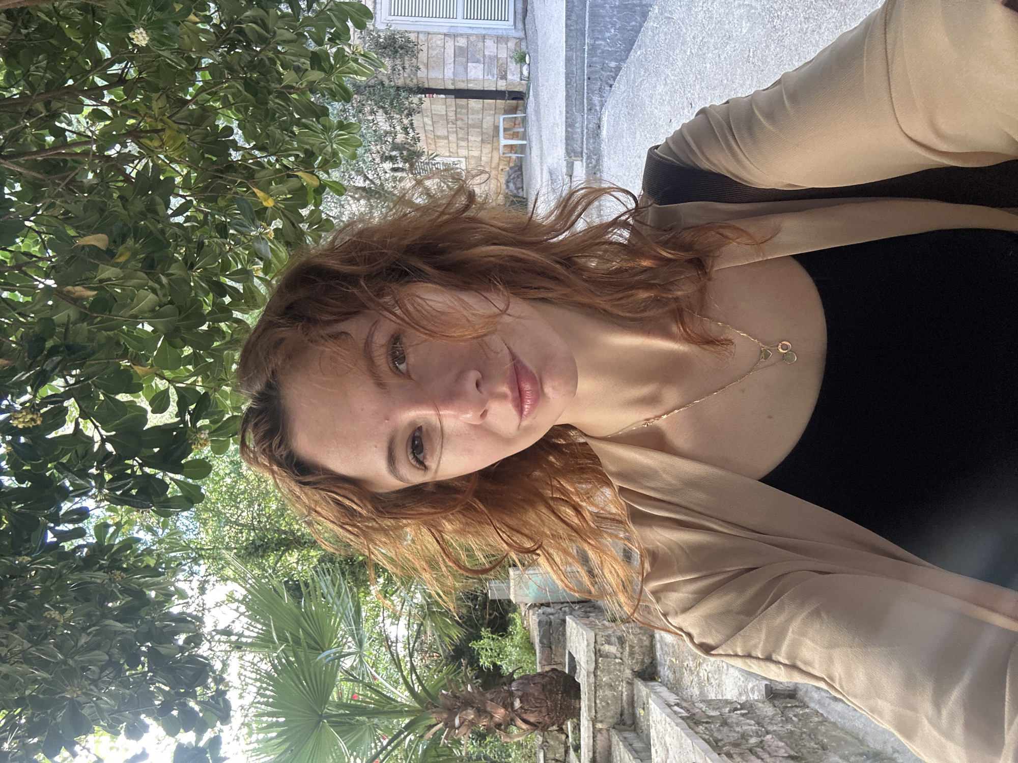 A selfie photograph presents a woman with red hair wearing a black top and beige cardigan, accessorized with a gold necklace. The camera angle is low, framing her face centrally, with an outdoor backdrop that suggests she's in Petrovac Centar, Montenegro.
Behind her, lush greenery fills the frame, including tree foliage, a palm tree, and other vegetation, indicative of a park or garden setting. Elements of built structure are also visible, which include a stone wall with steps, indicative of architectural elements that are characteristic of locations with similar surroundings, like Petrovac Centar, Montenegro.
The picture appears to have been taken during daylight hours.