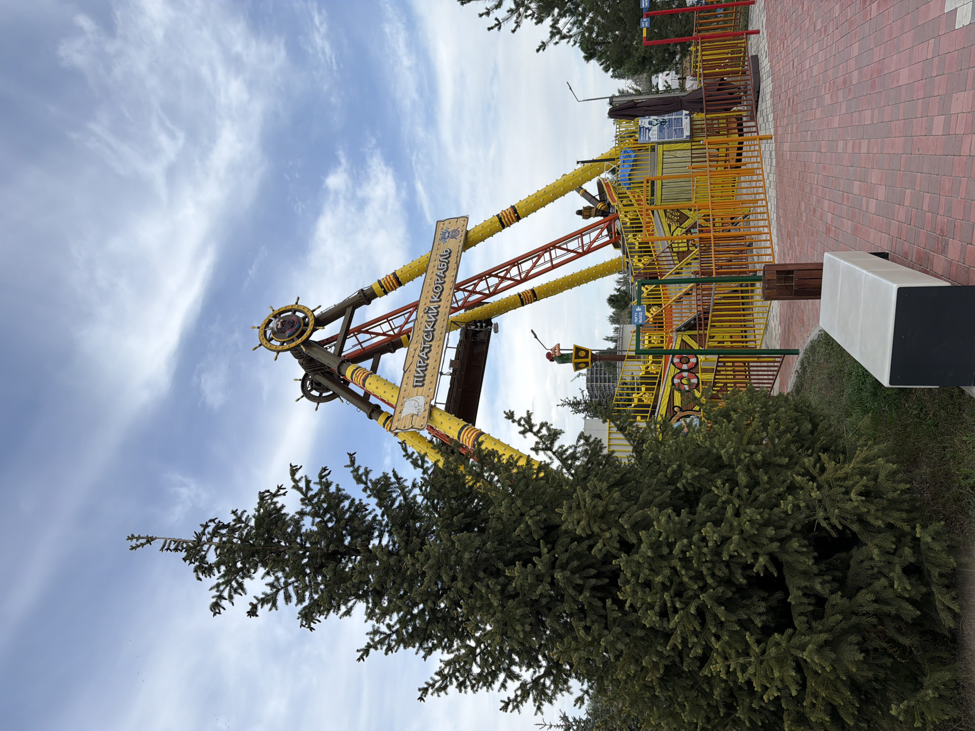 An amusement park ride, identified by a banner as "PIRATSKI KORABL" (Pirate Ship), is a prominent feature in the image. The ride is a large pendulum swing with a pirate ship theme, painted in yellow and red. It is set against a backdrop of a cloudy blue sky. In the foreground, a tall evergreen tree stands to the left, with a paved walkway and a bench to its right. The surrounding area appears to be an outdoor recreational space, possibly part of a larger park or city, with other structures and fencing visible in the background. The scene is captured during daylight hours.