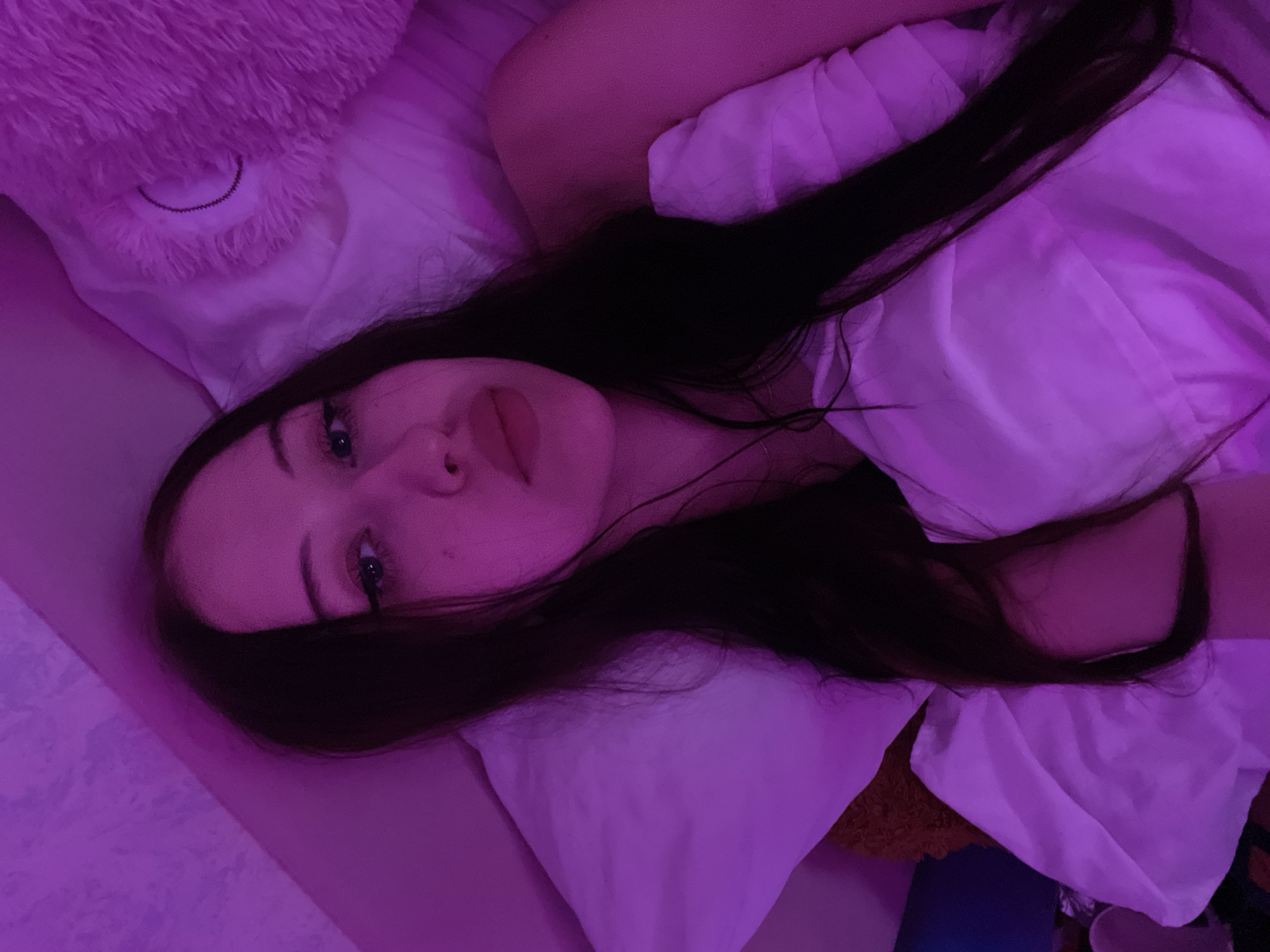 A young woman with long, dark hair lies in a bed, illuminated by purple lighting. She is seen from a high angle, with her head resting on a white pillow. Her face is partially obscured by her hair, but her eyes, nose, and lips are visible. She appears to be looking towards the camera. The background includes a fluffy pink object, possibly a stuffed animal, and a white duvet. The scene suggests a bedroom setting. There is no information in the image that places the location in Votkinsk, Russia.