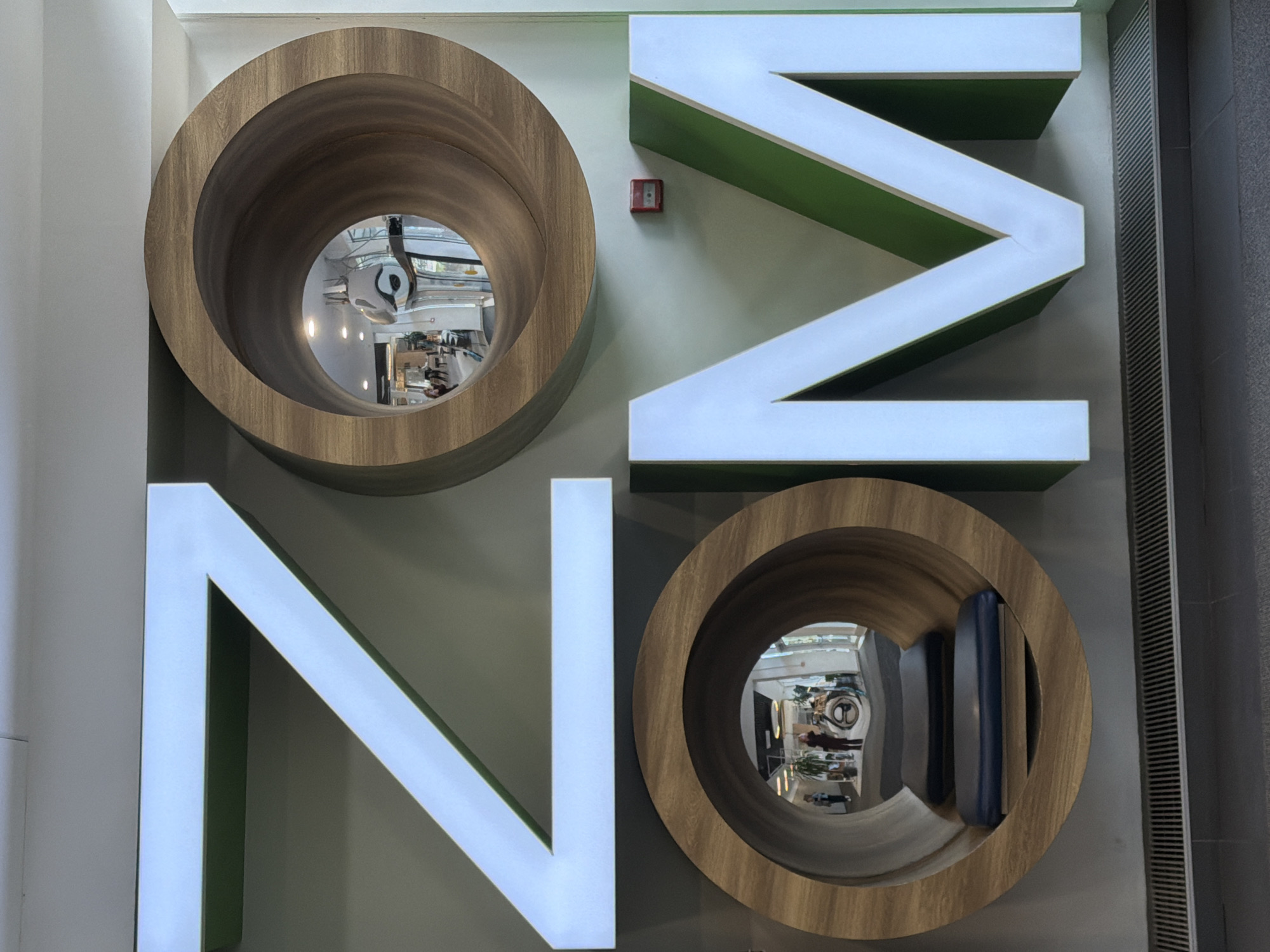The image shows a sign that spells "NO" vertically. The letters "O" are wooden circles with mirrored centers. The letter "N" is made of white blocks with green insets. The sign is on a light gray wall. A red fire alarm box is visible between the two letters. A vertical air conditioning vent is on the right side. Based on the reflection in the mirrored "O" letters, it appears to be inside a store or business. It is located in Saint Petersburg, Russia.