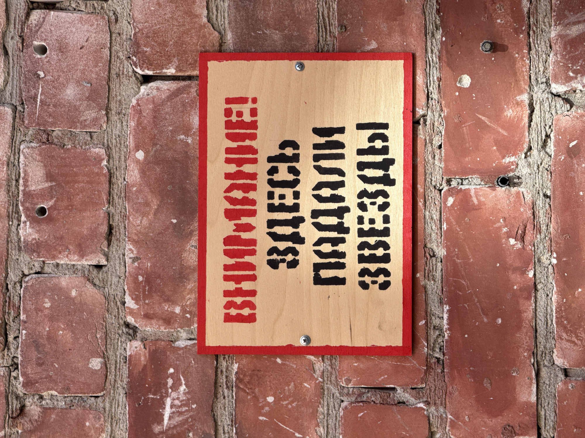 The image shows a sign attached to a red brick wall. The sign is rectangular with a red border and a light-colored wooden interior. It has text stenciled on it in red and black. The red text at the top reads "ВНИМАНИЕ!" (Attention!). Below that, in black, it reads "ЗДЕСЬ" (Here), then "ПАДАЛИ" (fell), and finally "ЗВЕЗДЫ" (stars). This translates to "ATTENTION! STARS FELL HERE". The sign is secured to the wall with two screws. The brick wall appears aged and weathered, with visible mortar lines and some imperfections on the bricks. The overall environment suggests an outdoor setting. The sign appears to be a novelty or humorous warning, possibly related to an event or legend. Without further context, it is impossible to definitively determine the exact location in Nizhny Novgorod, Russia, but it is likely a public space where such a sign would be placed.