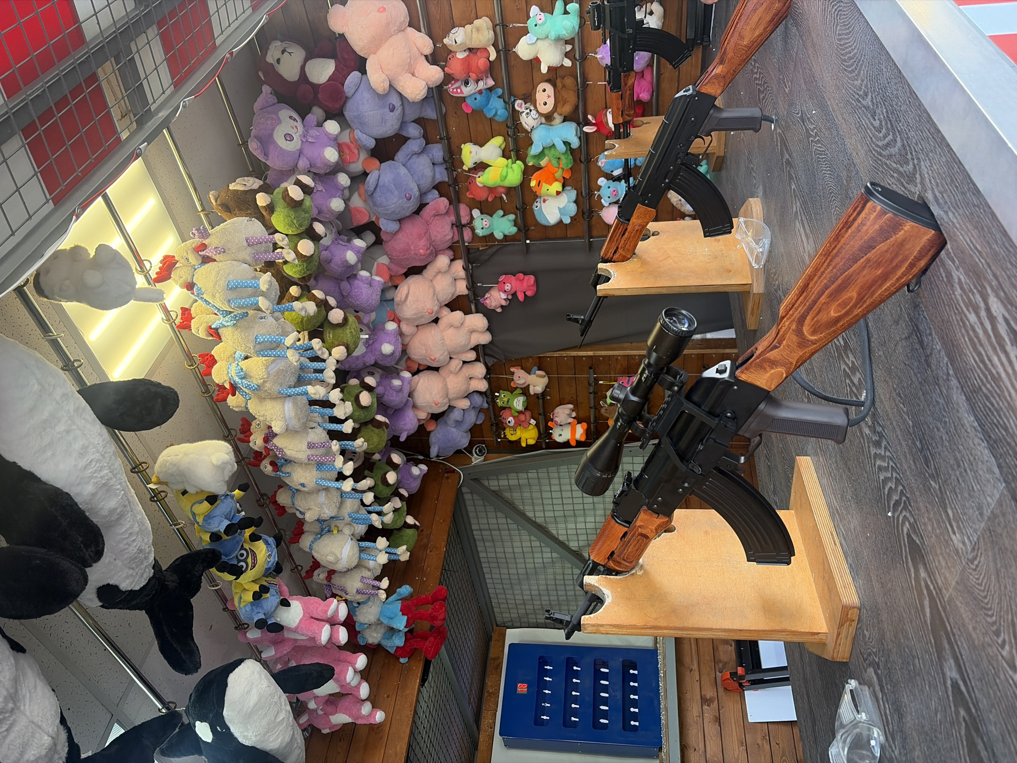 This image captures an indoor carnival or amusement park shooting game booth, likely located in Saint Petersburg, Russia. The scene is dominated by a display of numerous plush toys and two rifle-like objects, which appear to be airsoft guns or similar devices used for target practice.
On the right side of the frame, two long guns with rich brown wooden stocks and black barrels are mounted on separate wooden shelves, angled upwards. The lower rifle is equipped with a black scope, suggesting a game of precision. The guns are positioned for players to aim towards targets.
To the left and stretching across the background, a vast array of plush toys is visible, hanging from hooks and shelves. These prizes come in various shapes, sizes, and colors, including large white and black orca-like toys, yellow Minions, purple monsters, pink pigs, brown monkeys, and various other animals resembling chickens or sheep. They are densely packed, creating a vibrant and appealing display for potential winners.
At the bottom center, a blue rectangular box with multiple small white inserts or sensors is visible. This likely serves as the target system for the shooting game. A small, unidentifiable red logo or symbol is present on this blue box. In the upper left, a red and white metal cage-like structure suggests the boundary of the game booth. The overall setup indicates an interactive activity where players use the rifles to hit targets and win the displayed plush prizes. The lighting is artificial, common for an indoor setting. No specific human activity is depicted, but the arrangement clearly implies a game awaiting players.