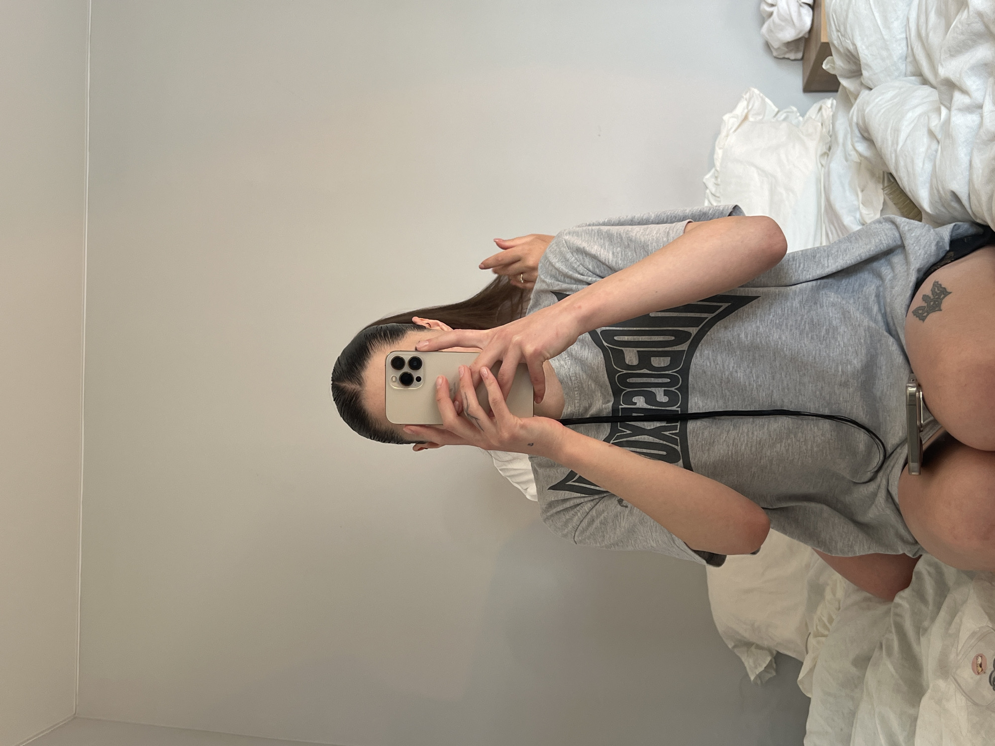 A person takes a mirror selfie with an iPhone while sitting on a bed covered in white linens. The person is wearing a gray t-shirt with a black design. They have long, dark hair pulled back and a tattoo on their left thigh. The background is a plain, light gray wall. There is no information available to determine the location is Ternopil, Ukraine.