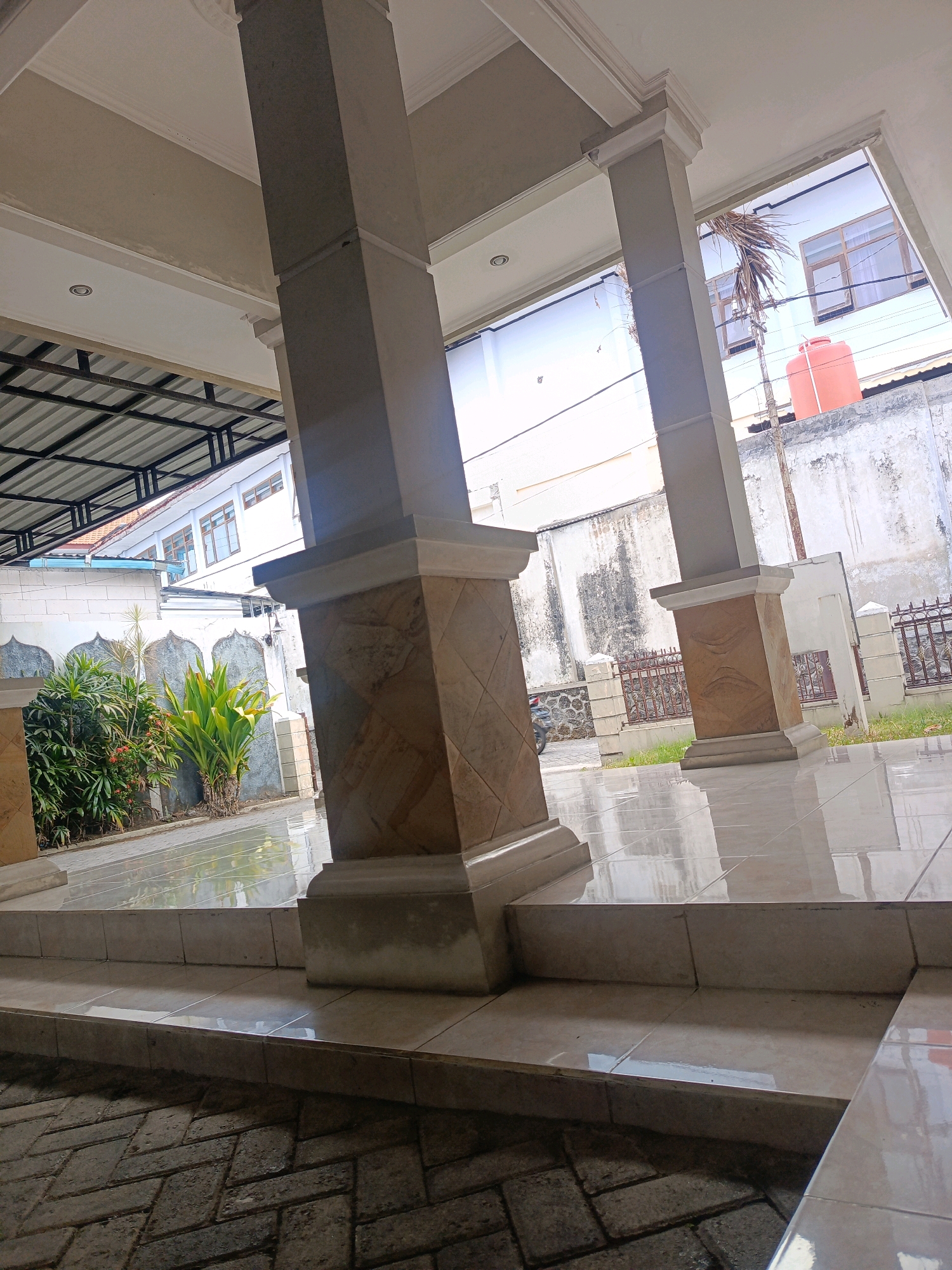 The image captures a courtyard or porch area, likely part of a building, possibly a mosque or a public facility in Kebonagung, Indonesia. Two large, square pillars with decorative bases dominate the foreground, angled to create a sense of depth. The pillars are constructed of light-colored stone or concrete, with the lower portions featuring a patterned tile. Beyond the pillars, a tiled floor reflects the bright daylight. To the left, lush green plants are visible, including some with broad, tropical leaves and smaller flowering bushes. In the background, a white building with multiple windows stands behind a decorative stone wall with arched elements. A bright orange water tank is situated on top of the wall. A corrugated metal roof structure is partially visible on the far left, suggesting an awning or covered area. The ground in front of the tiled steps is paved with dark, interlocking bricks laid in a herringbone pattern. The overall scene is one of a well-maintained outdoor space, bathed in bright sunlight, with a tropical atmosphere. There are no people or animals visible in the image.