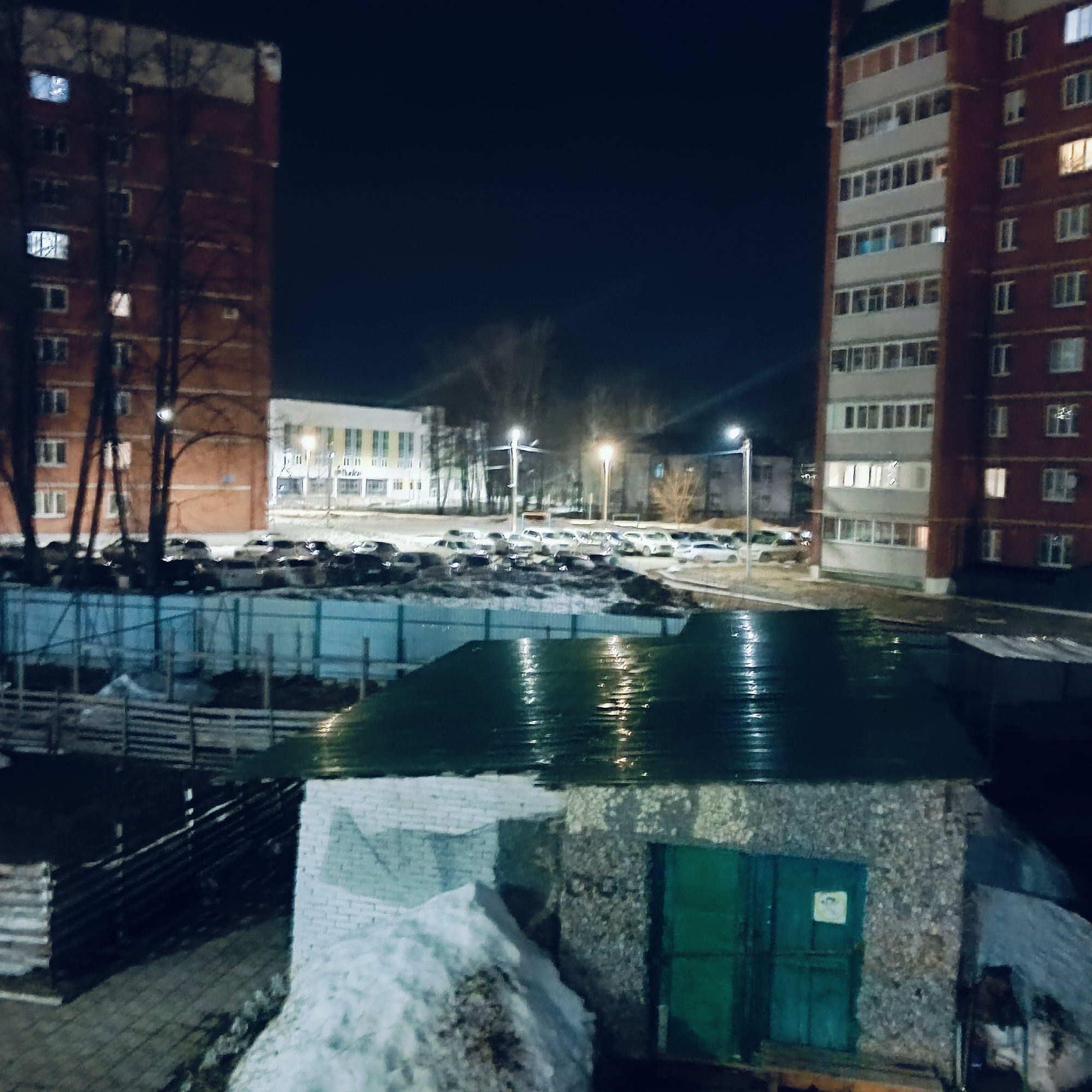 The image, taken at night in what is believed to be Chaikovsky, Russia, presents an urban scene dominated by multi-story residential buildings. The building on the left is a brick-faced structure with visible windows and dark tree silhouettes partially obscuring its facade. A building with a lighter, likely brick facade, stands to the right with more windows visible, some illuminated from within.
Between the two buildings is an illuminated parking area filled with numerous cars. Streetlights cast a glow over this space, suggesting public lighting. In the foreground is a small building or shed with a green corrugated metal roof and a white-painted lower section. A pile of snow sits next to it. A wooden fence can be seen behind the shed.
The scene lacks observable human activity, focusing instead on the urban infrastructure under the illumination of the night.