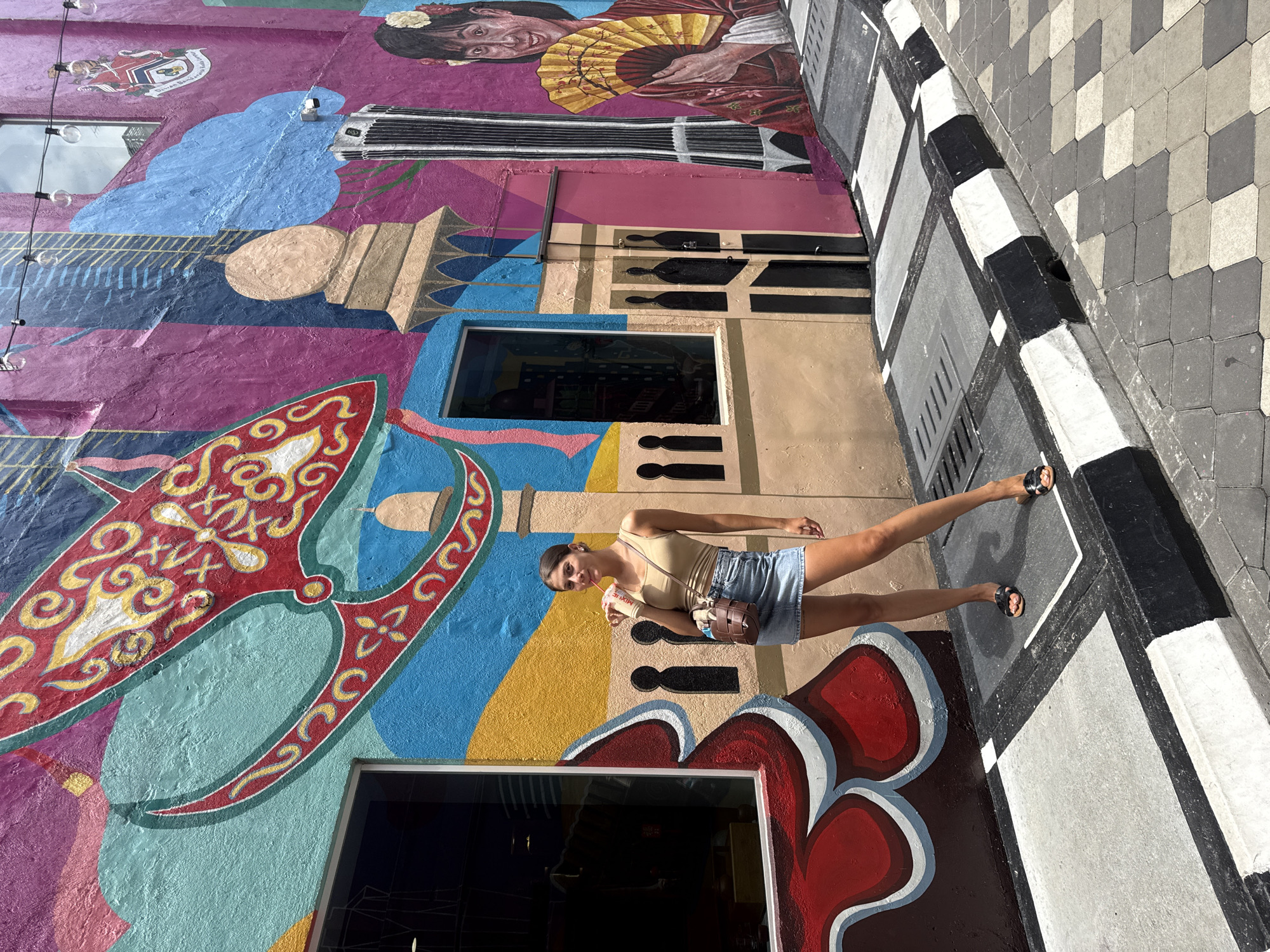 A young woman stands on a black and white patterned street beside a vibrant mural in Kuala Lumpur, Malaysia. The mural features a diverse mix of cultural and artistic elements, including portraits of people, architectural motifs, and abstract designs in bright colors. The woman is wearing a light-colored top, denim shorts, and sandals, holding a cup with a straw in one hand. She appears to be posing for a photograph or enjoying the artwork. The street is paved with patterned tiles, and the curb is painted with alternating black and white stripes. Windows and reflections are visible within the mural itself, indicating the presence of buildings or establishments behind the artwork.