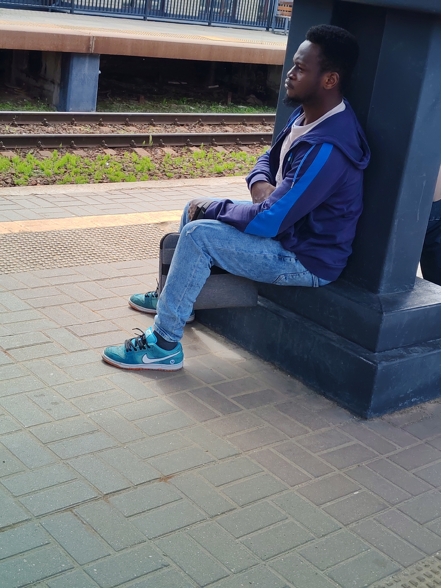 The image features a man of Black ethnicity sitting on a train platform during what appears to be daytime. He is depicted in profile, looking towards the left with a neutral or pensive expression. The man wears a dark blue hooded jacket with light blue stripes on the sleeves, a light-colored t-shirt, and light blue ripped jeans. His feet are adorned with distinctive teal or turquoise Nike sneakers, which have a white swoosh, black laces, and orange soles. A small "SB" logo is visible on the side of one shoe near the heel.
He is seated, leaning against a large, dark grey or black pillar-like structure that rises from the platform. His arms are crossed over his lap. The platform itself is paved with light grey rectangular bricks laid in a herringbone pattern. In the background, beyond the platform edge, a set of train tracks is visible, with green vegetation growing between the rails and on the ballast. Further back, there is another raised platform or structure, brown in color, supported by blue pillars. The overall scene suggests a waiting area at a train station. While not explicitly visible in the image, the provided context specifies the location as Moscow, Russia.